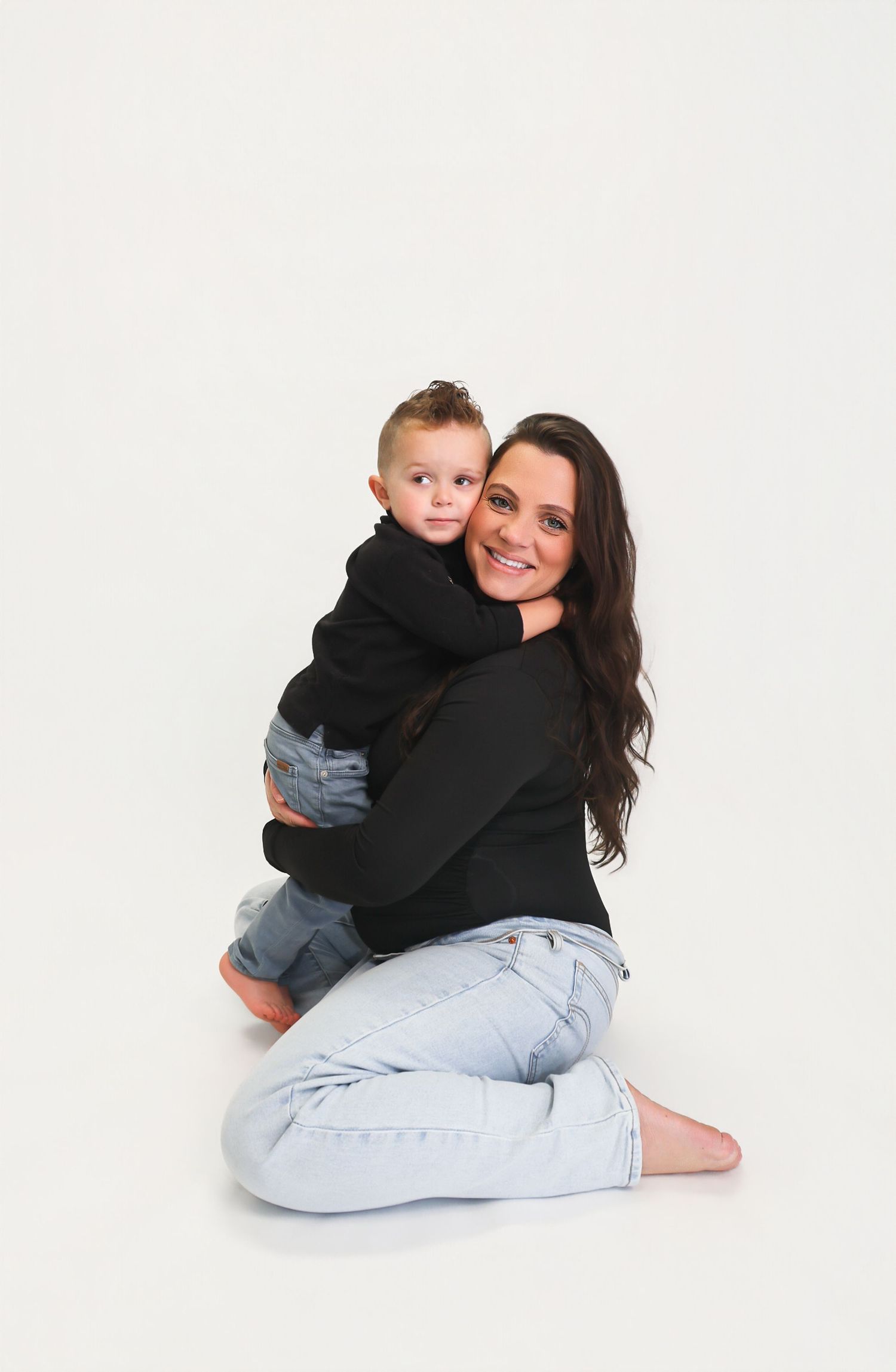 A woman with dark hair sits on the floor hugging a young boy, both wearing black tops and jeans, smiling warmly.