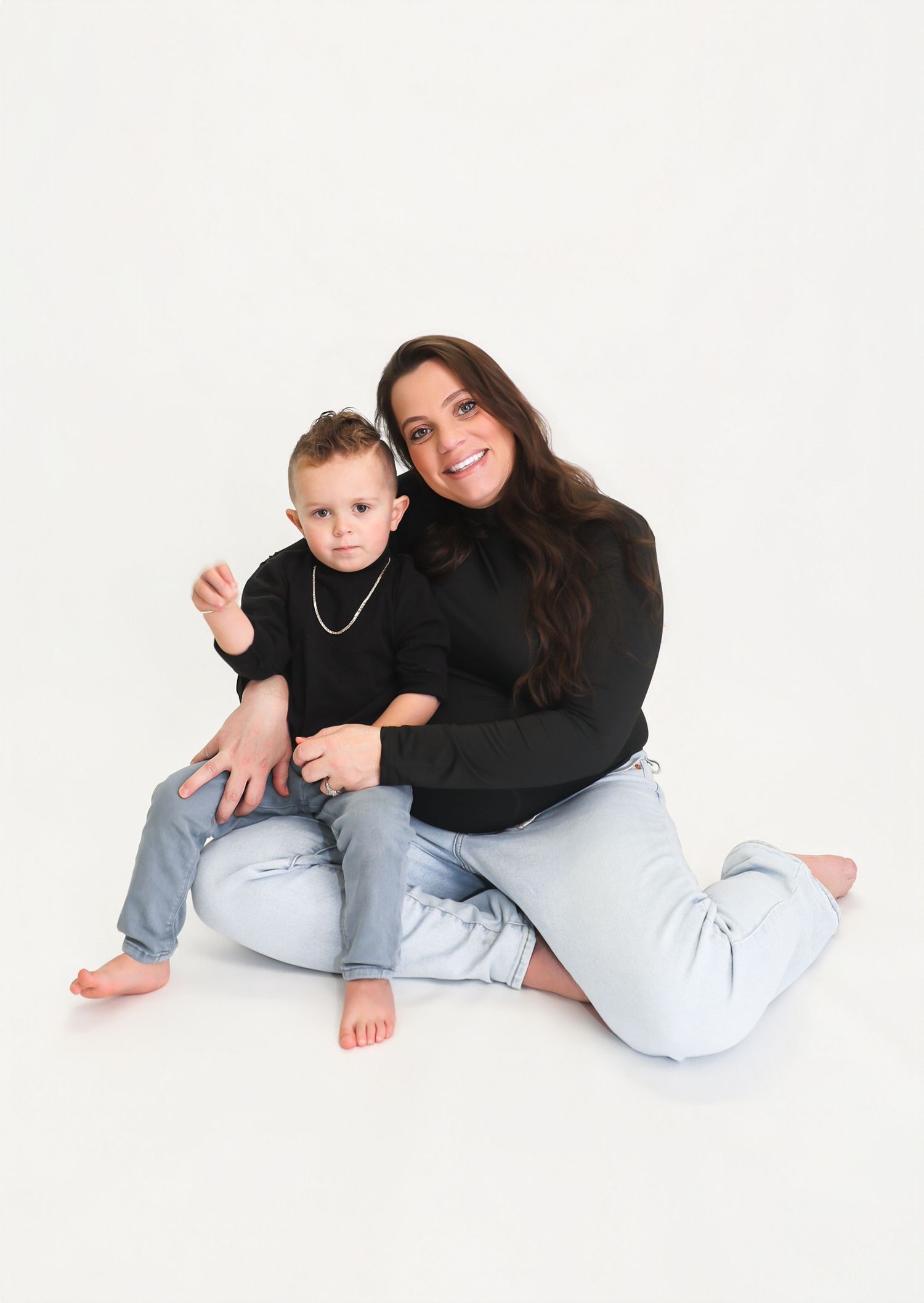 A woman and young boy in matching black tops and light jeans pose together on a white studio background.