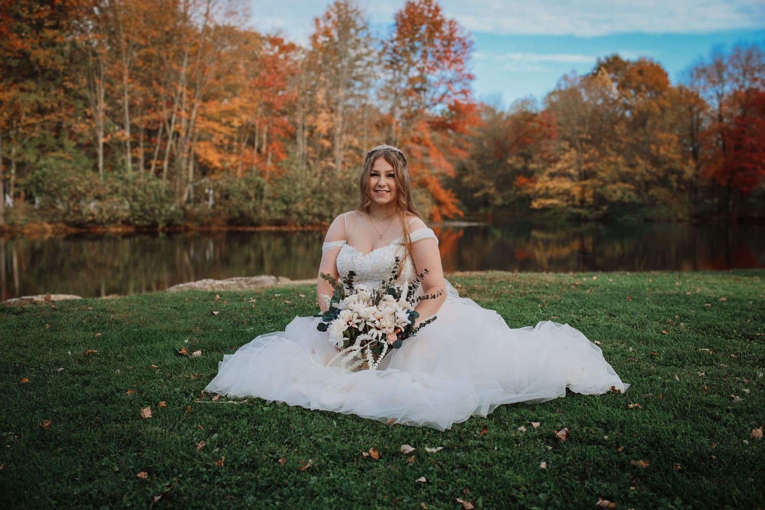 Bride in white gown sitting on grass with fall foliage and lake in background.