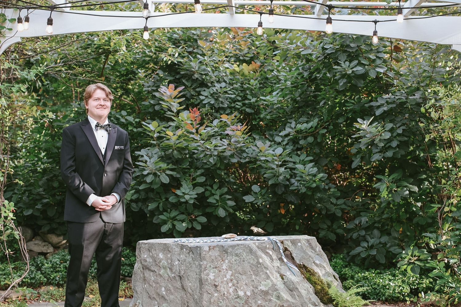 A wedding party poses outdoors in formal attire near a large stone wall surrounded by lush greenery.
