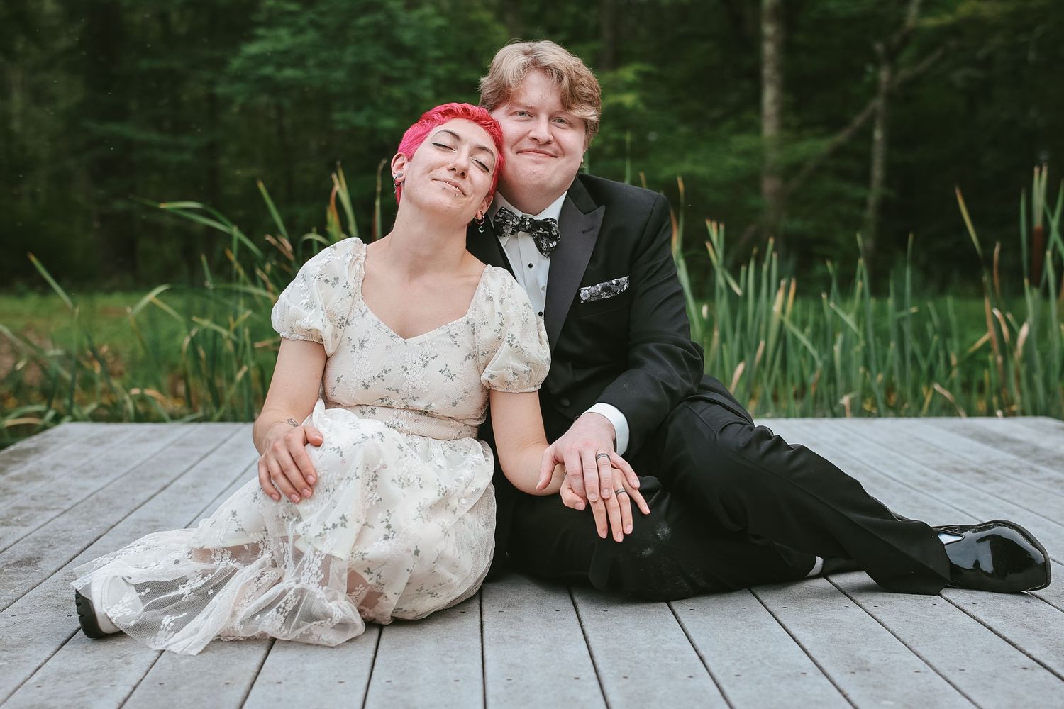 Couple in wedding attire sits and laughs together on wooden dock by a lake surrounded by greenery.