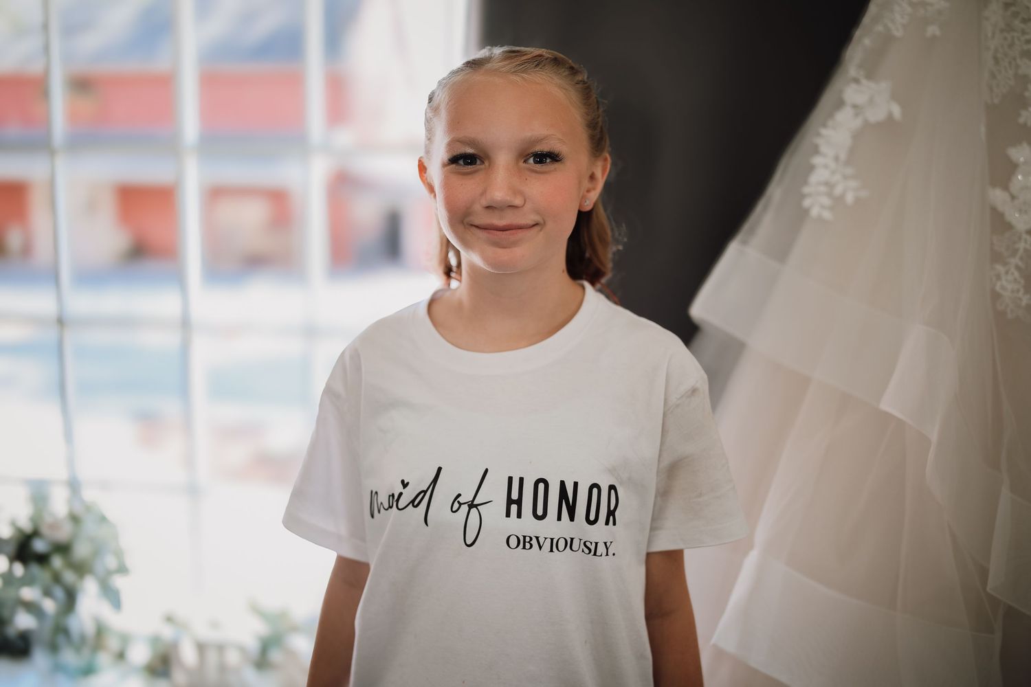 A young person wearing a white 'Maid of Honor' t-shirt smiles while standing next to a wedding dress.