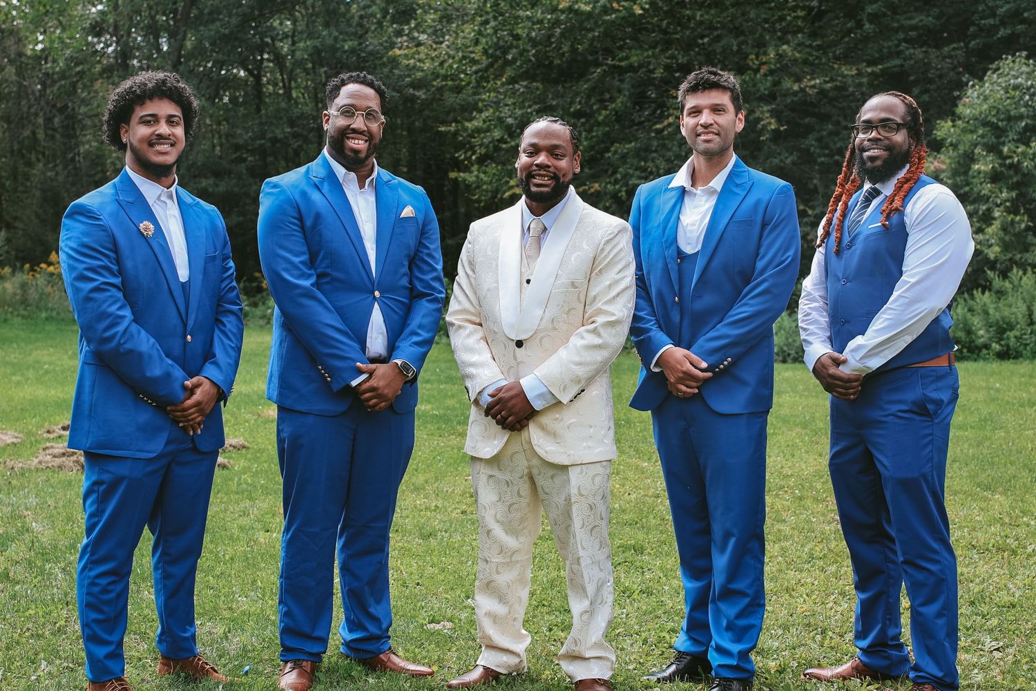 Groomsmen wearing bright blue suits pose with groom in white suit on grassy lawn during outdoor wedding.