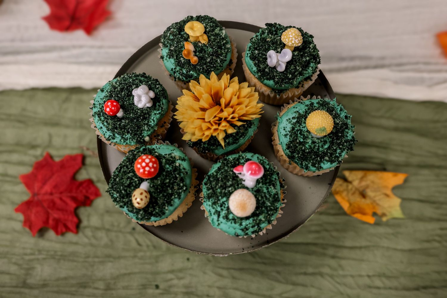Decorative cupcakes topped with green frosting, yellow flowers, and mushroom decorations arranged on a plate with fall leaves.