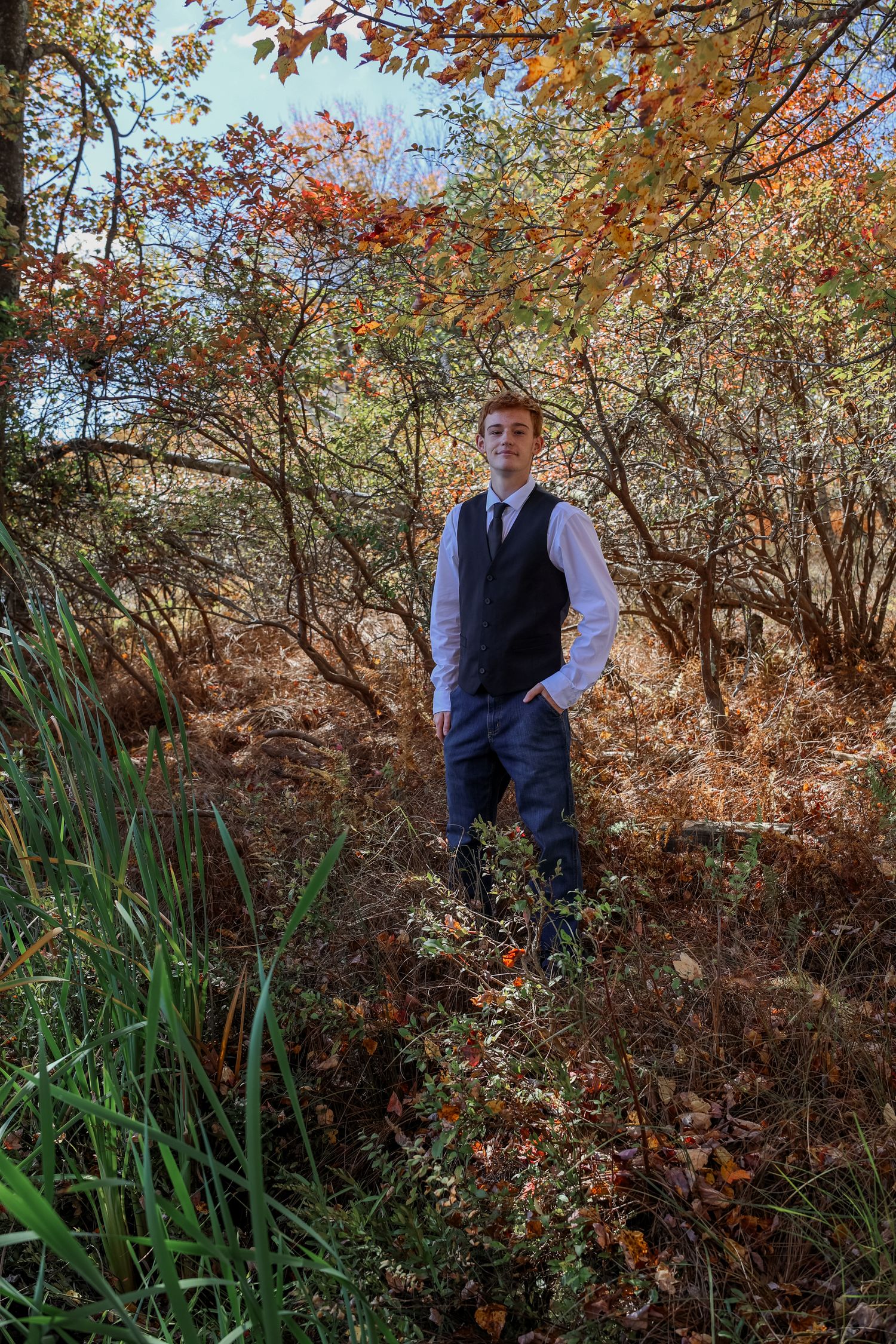 Fall foliage surrounds a figure in a navy vest and white shirt standing among autumn trees and tall grasses.