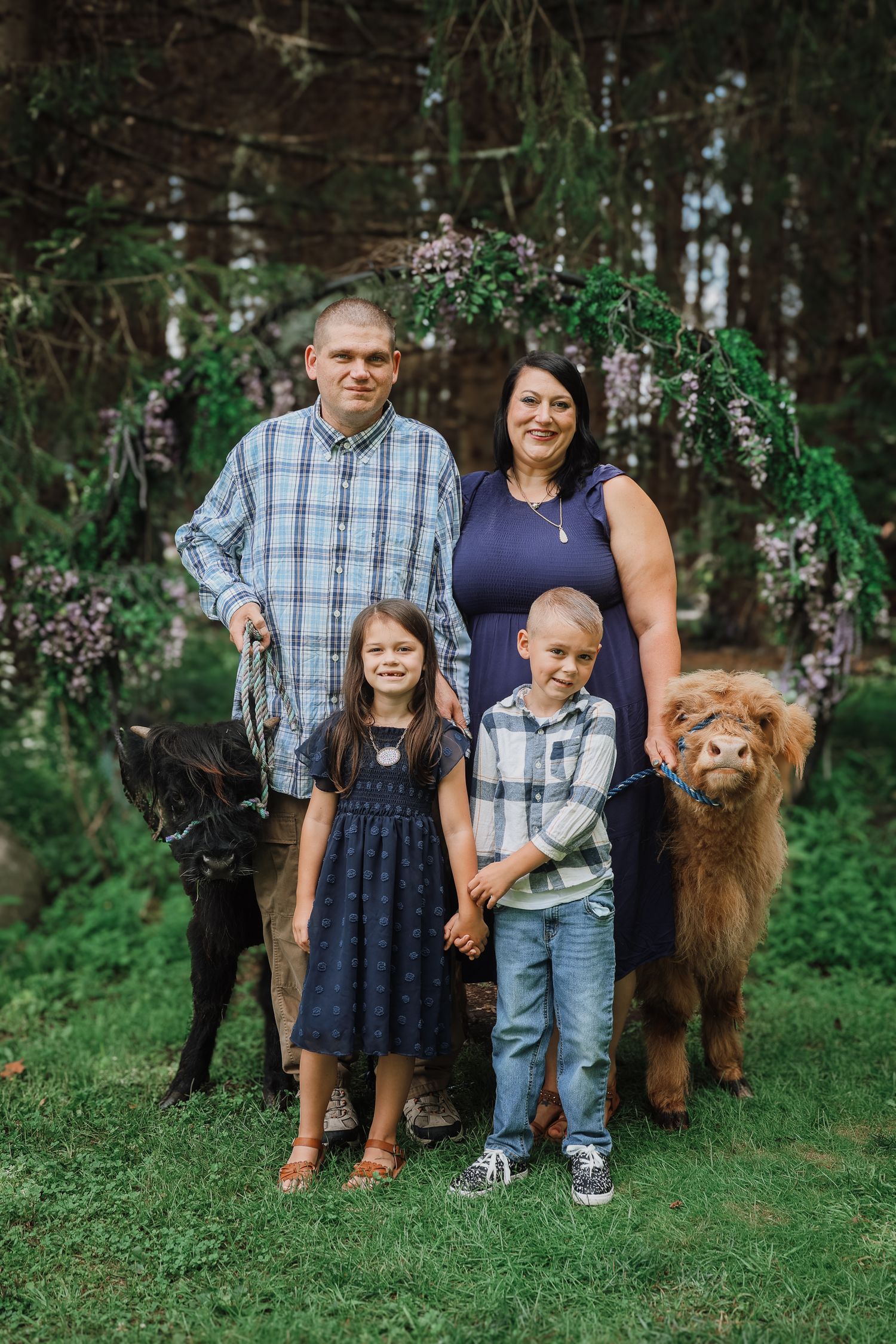 A family poses together with their fluffy brown dog on a grassy lawn surrounded by trees and flowering shrubs.
