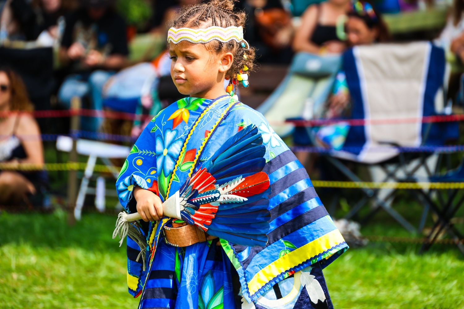 Young dancers in colorful tie-dye outfits perform together at an outdoor festival on a grassy field.