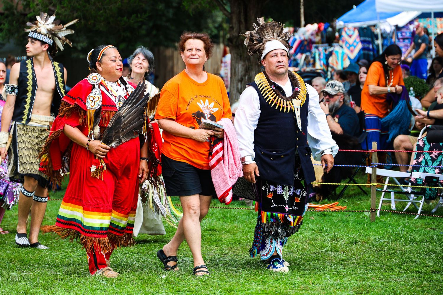 Dancers in traditional regalia and colorful outfits perform at an outdoor festival on a sunny day.