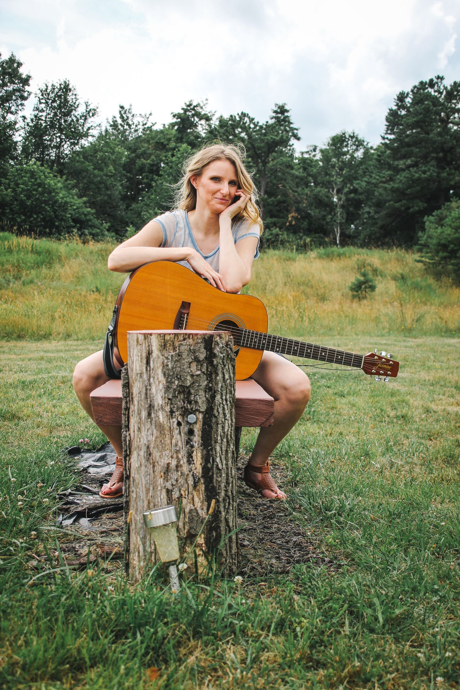 A musician sits on a tree stump in a grassy meadow playing an acoustic guitar on a summer afternoon.