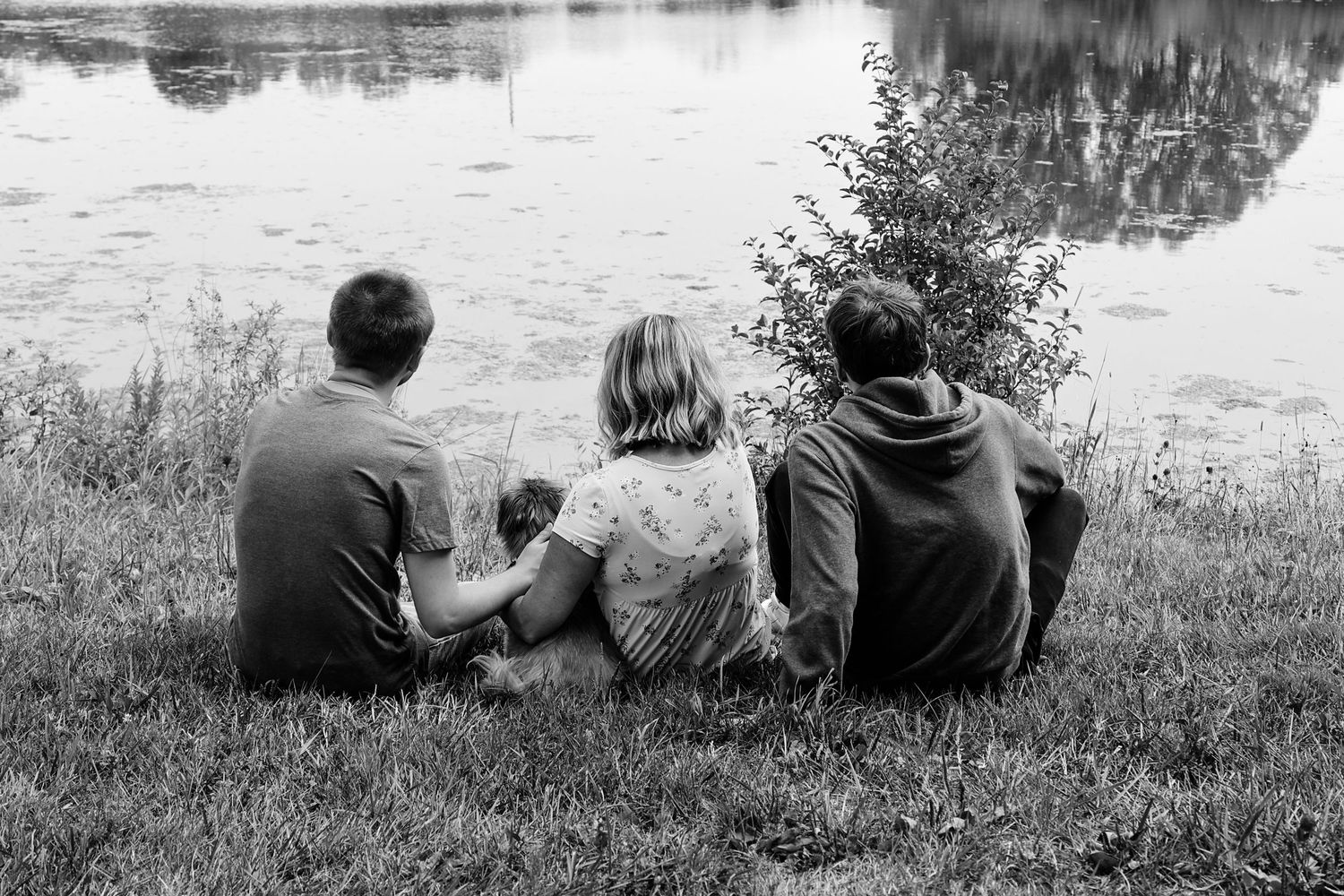 Two people sit together on a grassy riverbank in black and white, creating a peaceful scene of companionship.