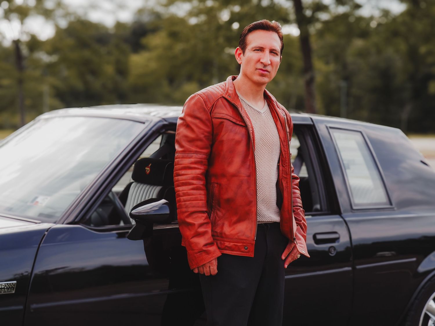 A person in a red leather jacket stands next to a black car in an outdoor setting.