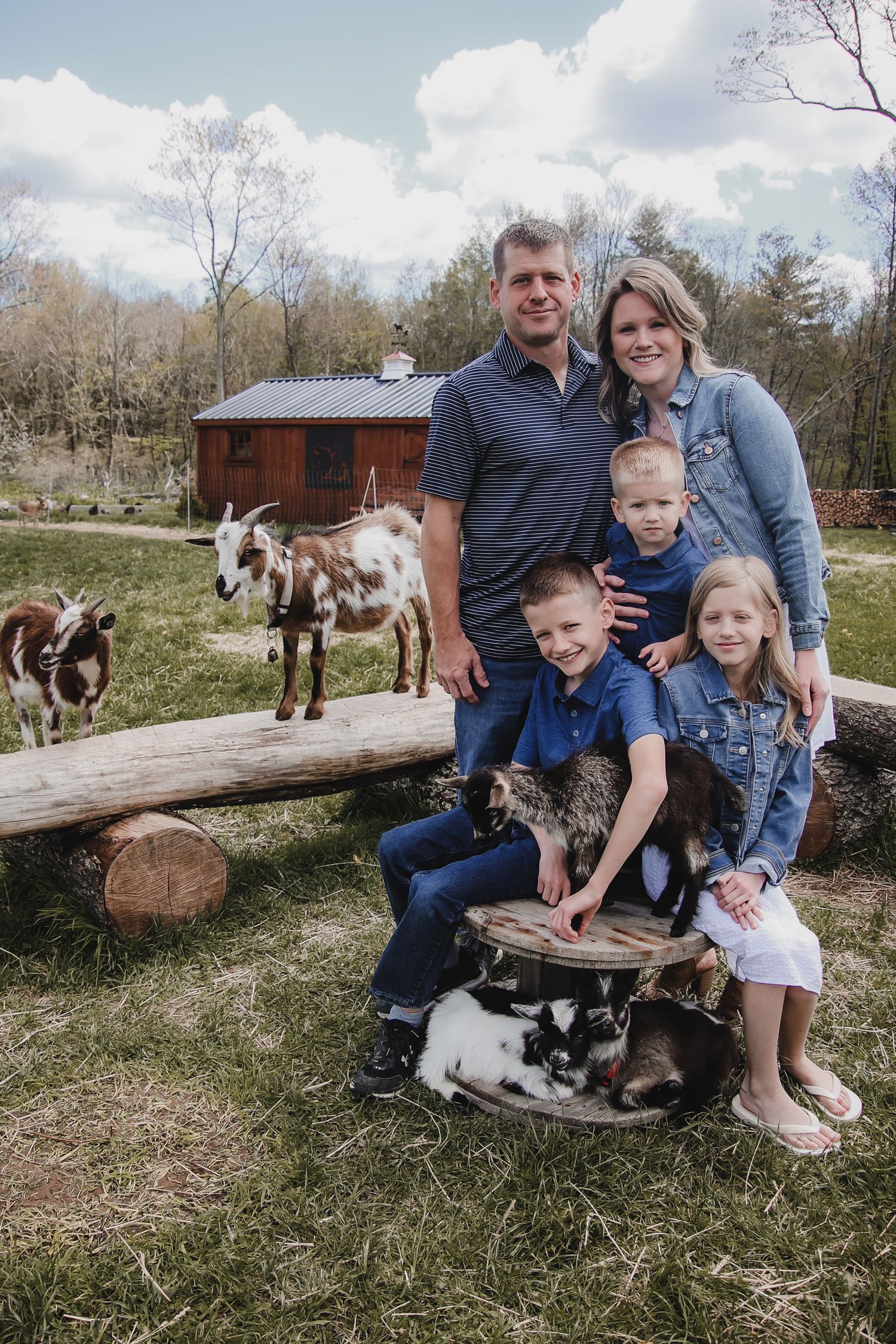 Family and children sitting together outdoors with horses and wooden logs in a grassy field on a sunny day.