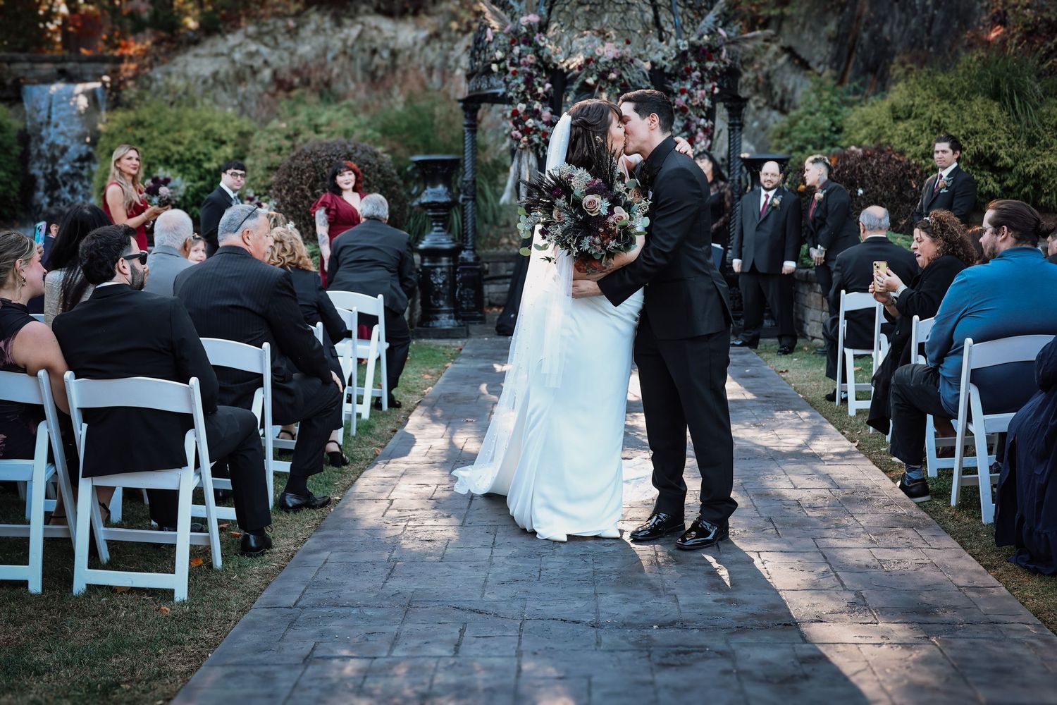 A wedding ceremony takes place outdoors with guests seated in white chairs along a garden pathway.