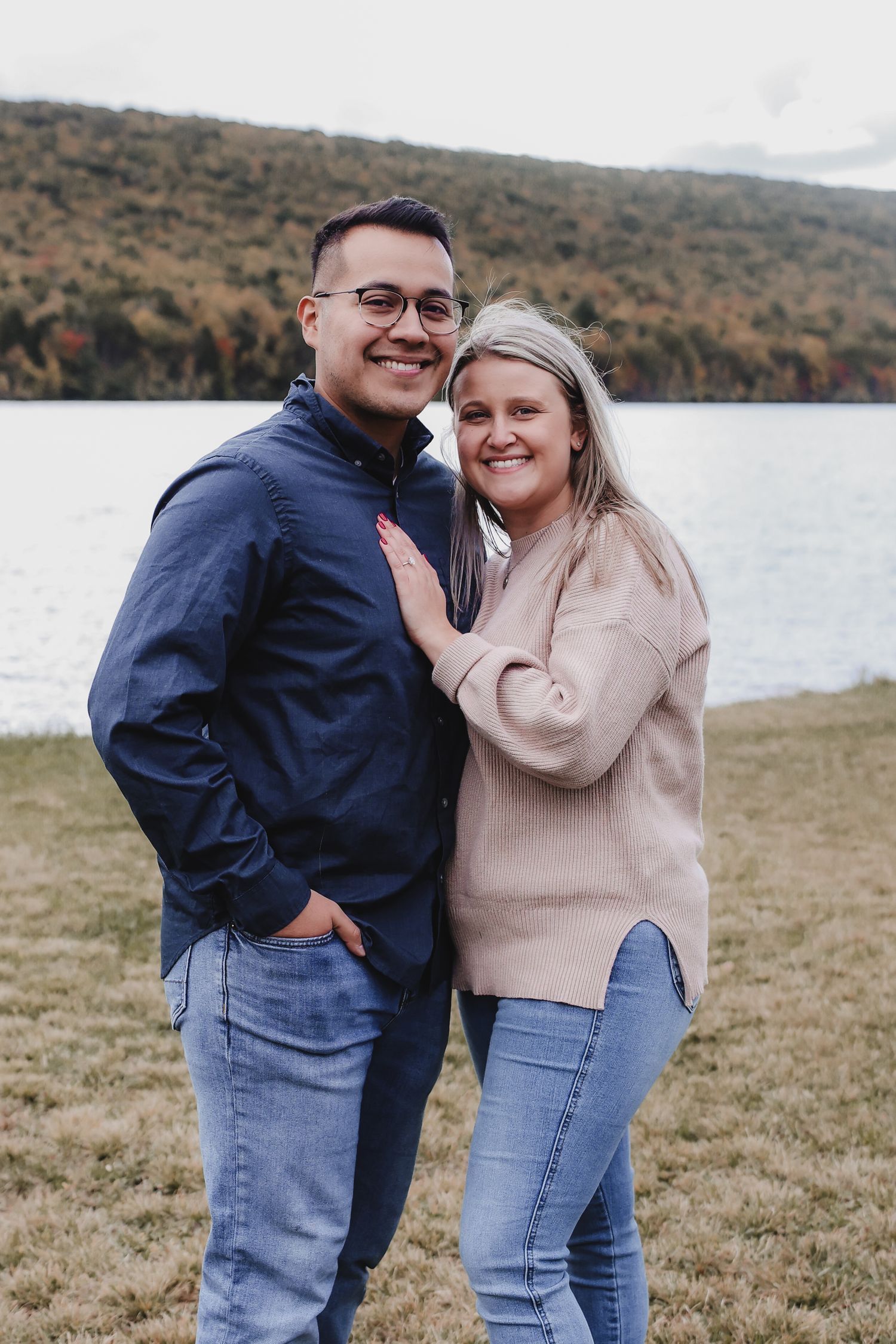 A couple embraces while standing on grassy shore of a lake with autumn hills in the background.