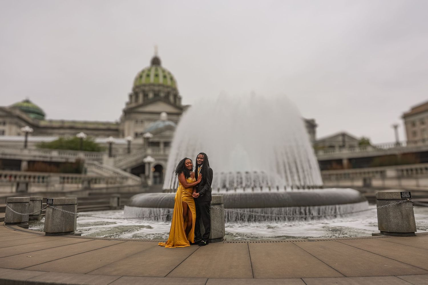 A series of water fountains spray in front of a historic building with a golden dome on a cloudy day.
