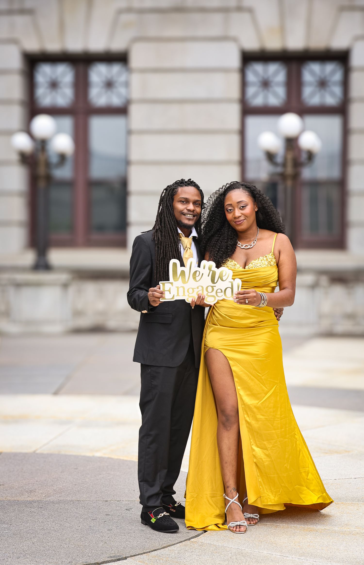 A couple poses together outdoors against a classic stone building, one in yellow dress and one in black attire.