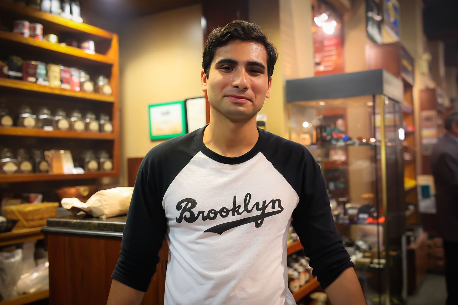 A person wearing a Brooklyn t-shirt stands in front of shelves stocked with various products in a retail store.