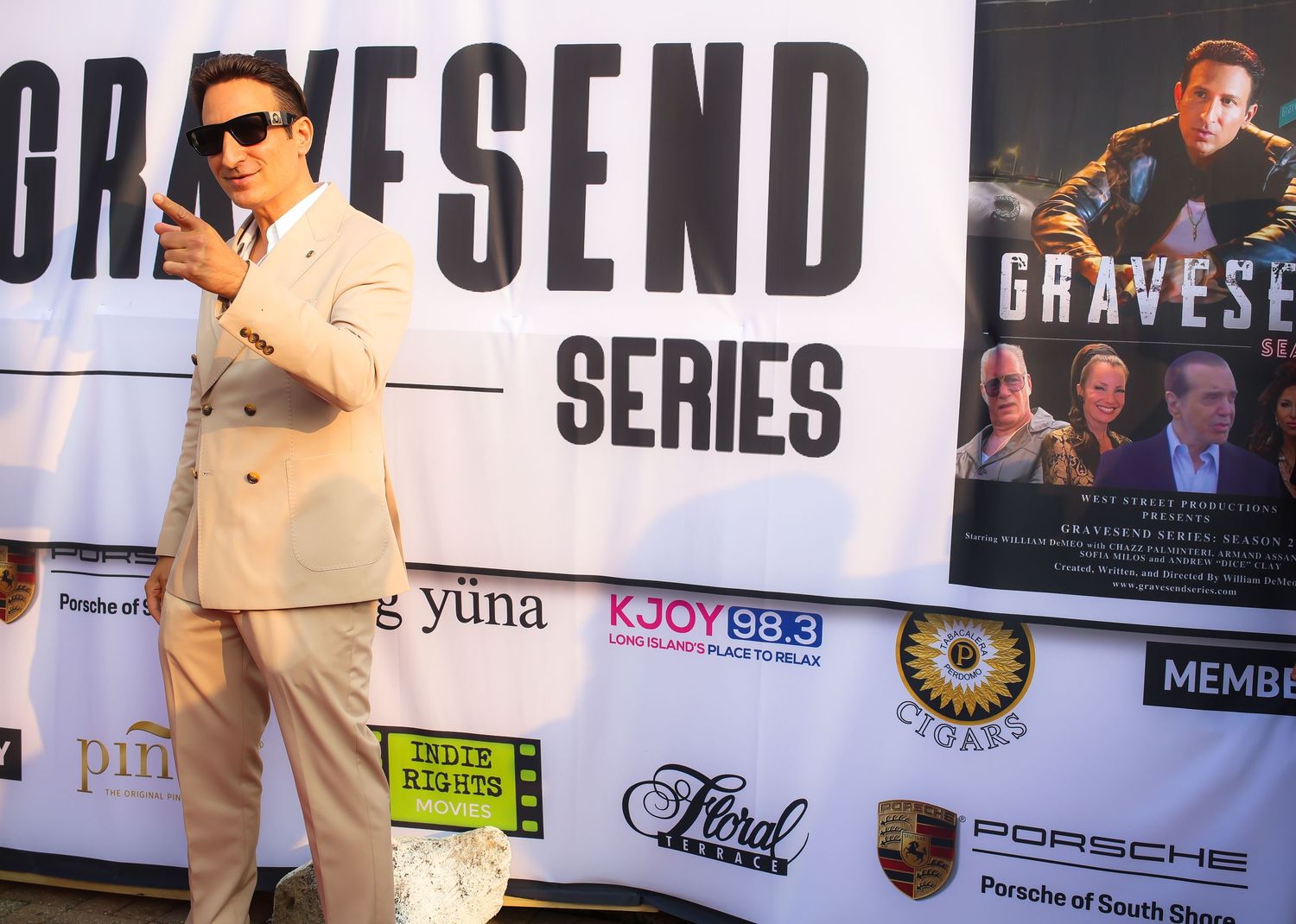 A person in a light colored suit poses in front of a Gravesend Series promotional backdrop at an event.