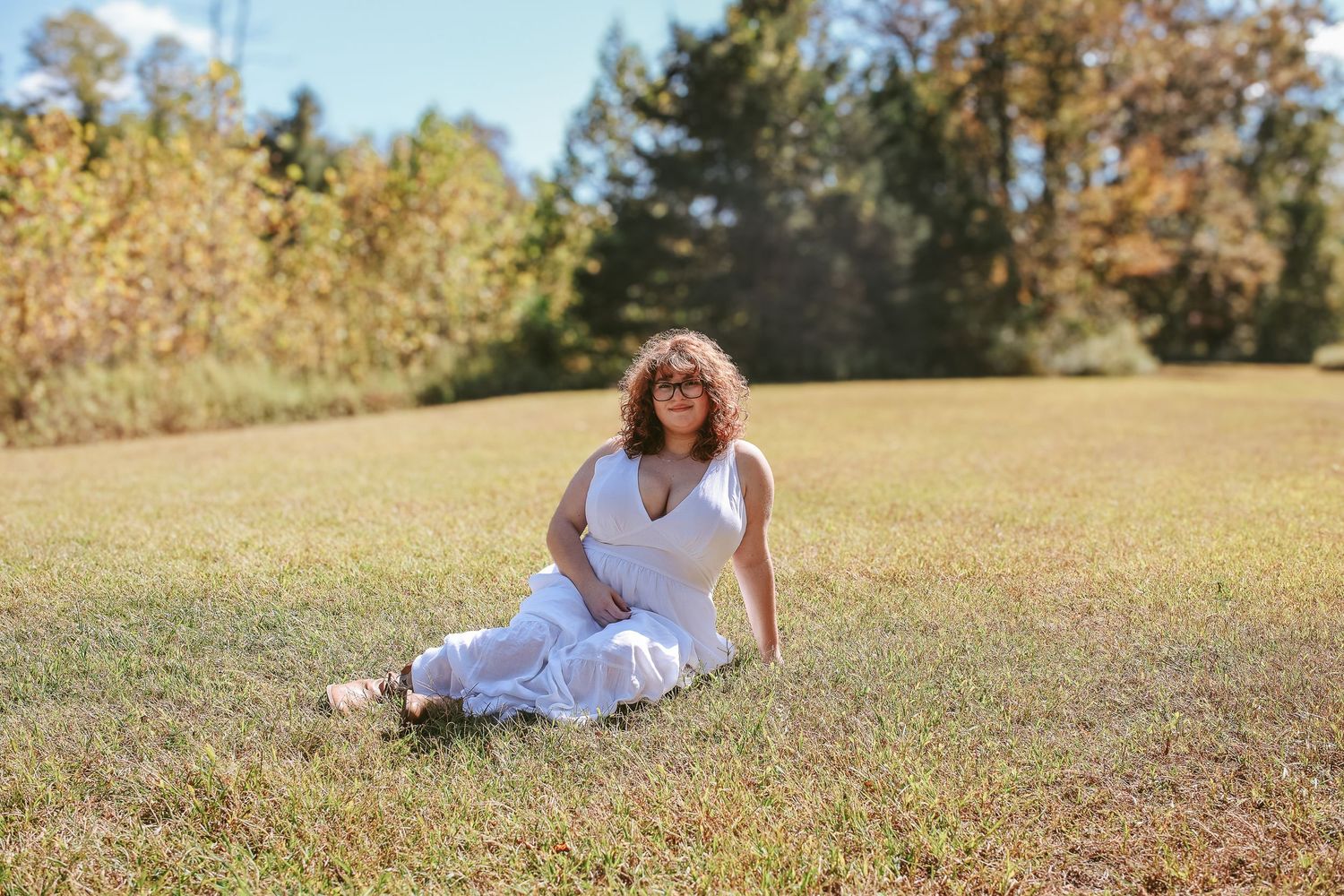 A person in a white dress lies on a grassy hill during a sunny autumn day with trees in the background.