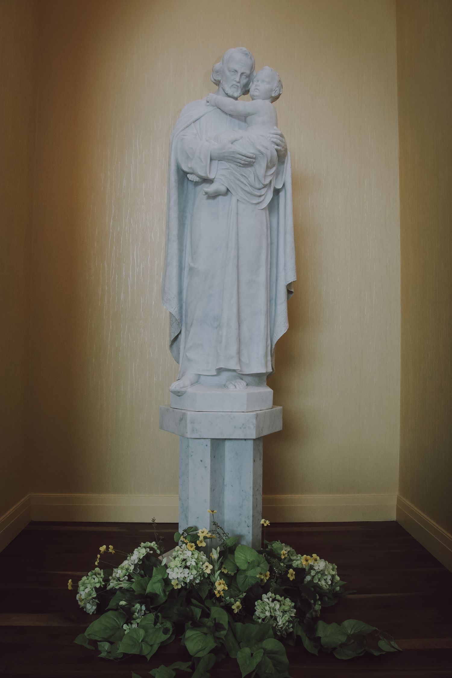 White marble statue of a robed figure stands against beige wall with green plants at its base.