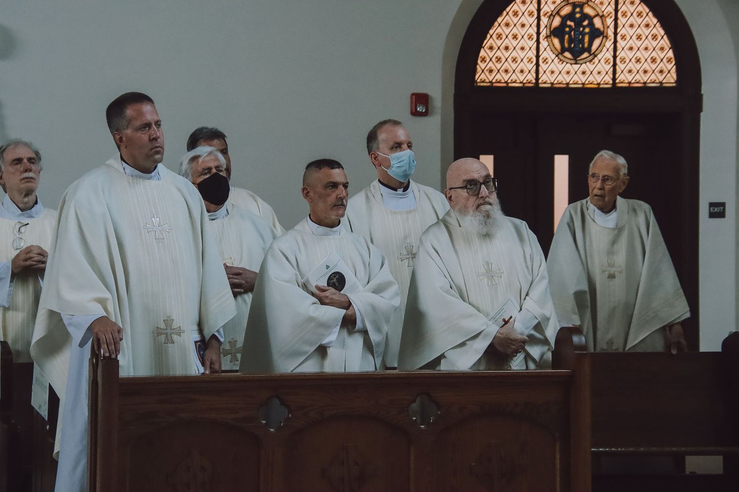 Clergy members wearing white robes gather in church pews during a religious service.