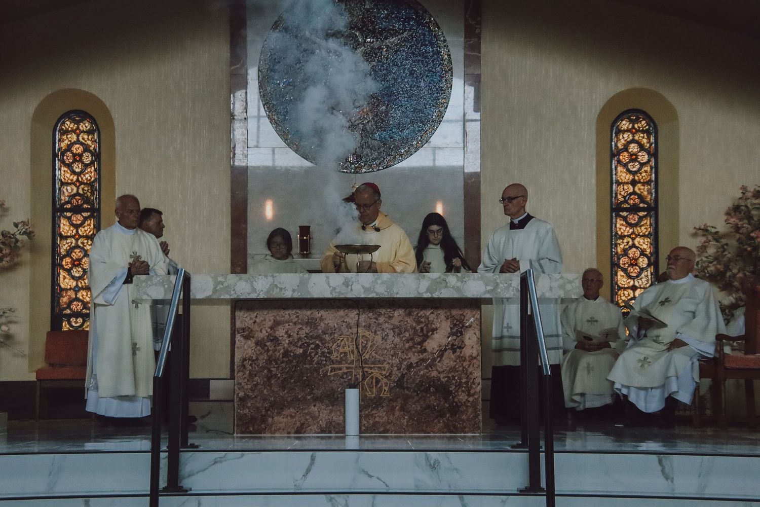 Religious ceremony taking place at an altar with stained glass windows and a large circular artwork in the background.