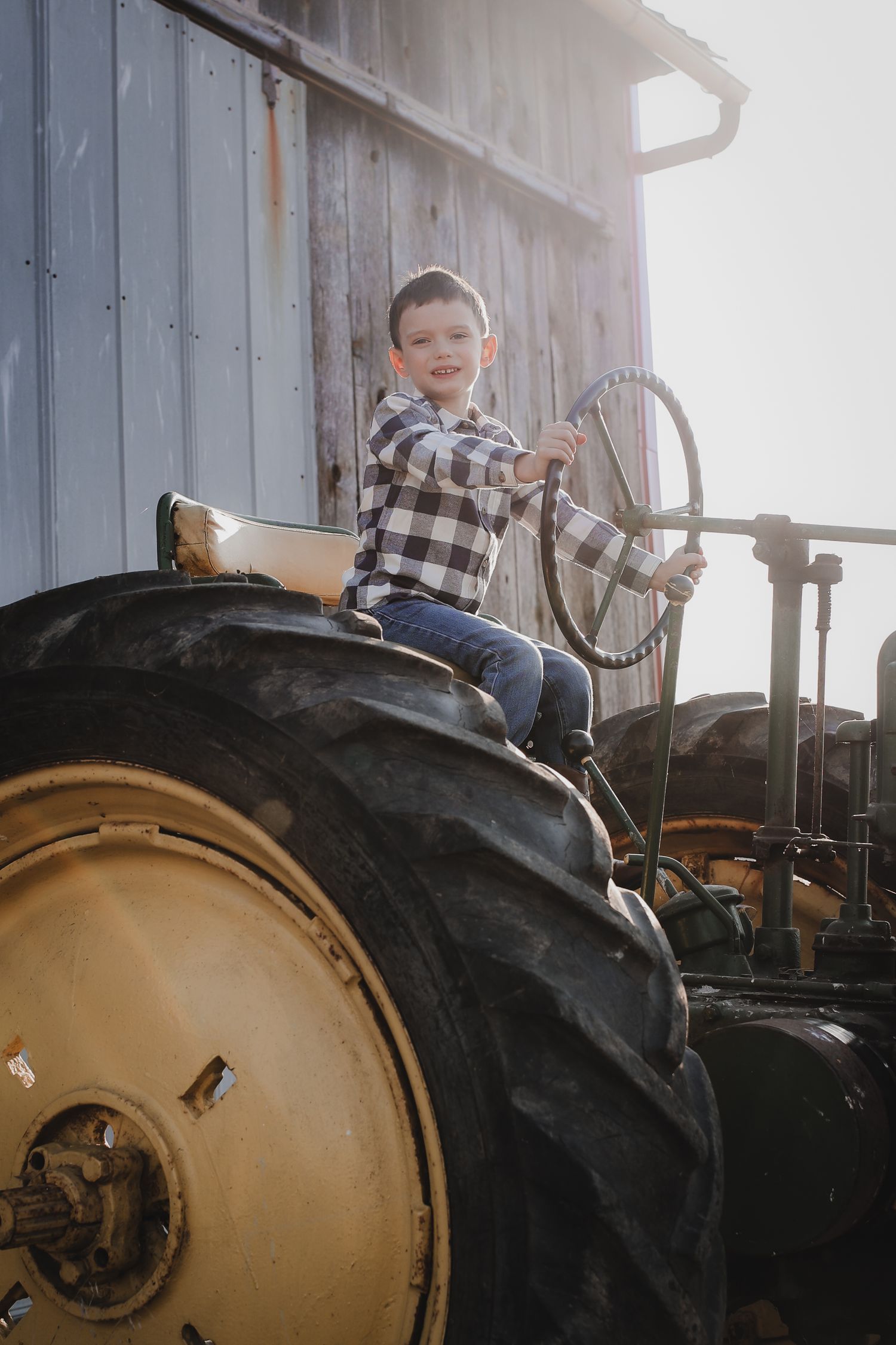 A young child sits atop a large vintage tractor wheel wearing a plaid shirt and jeans on a sunny day.