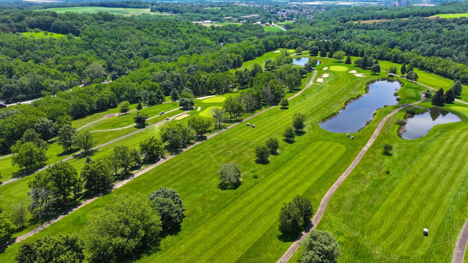 Aerial panoramic view of a lush green golf course with winding paths and trees nestled in rolling countryside landscape.