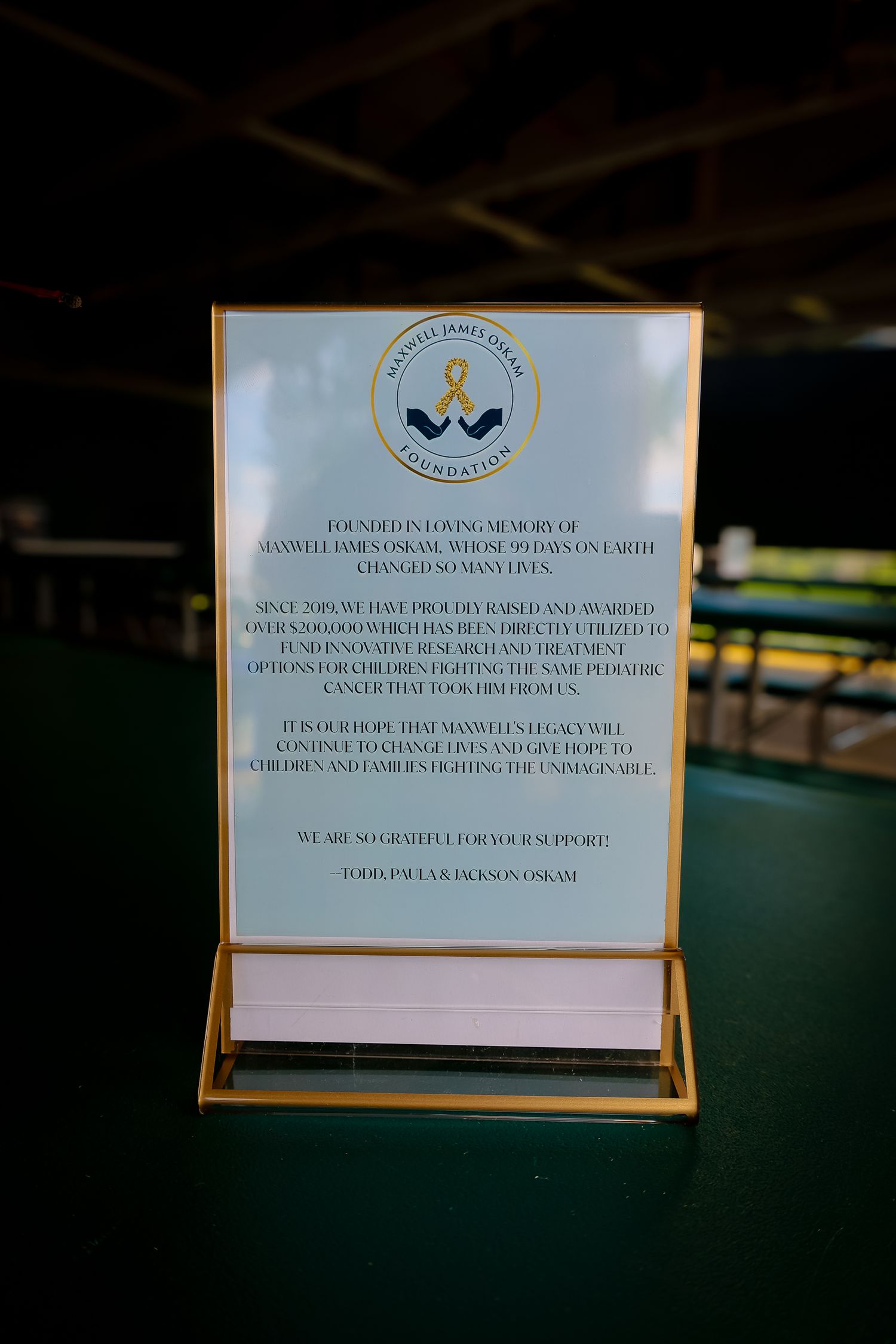 A framed sign with a logo and text displayed on a gold-trimmed stand against a dark background.