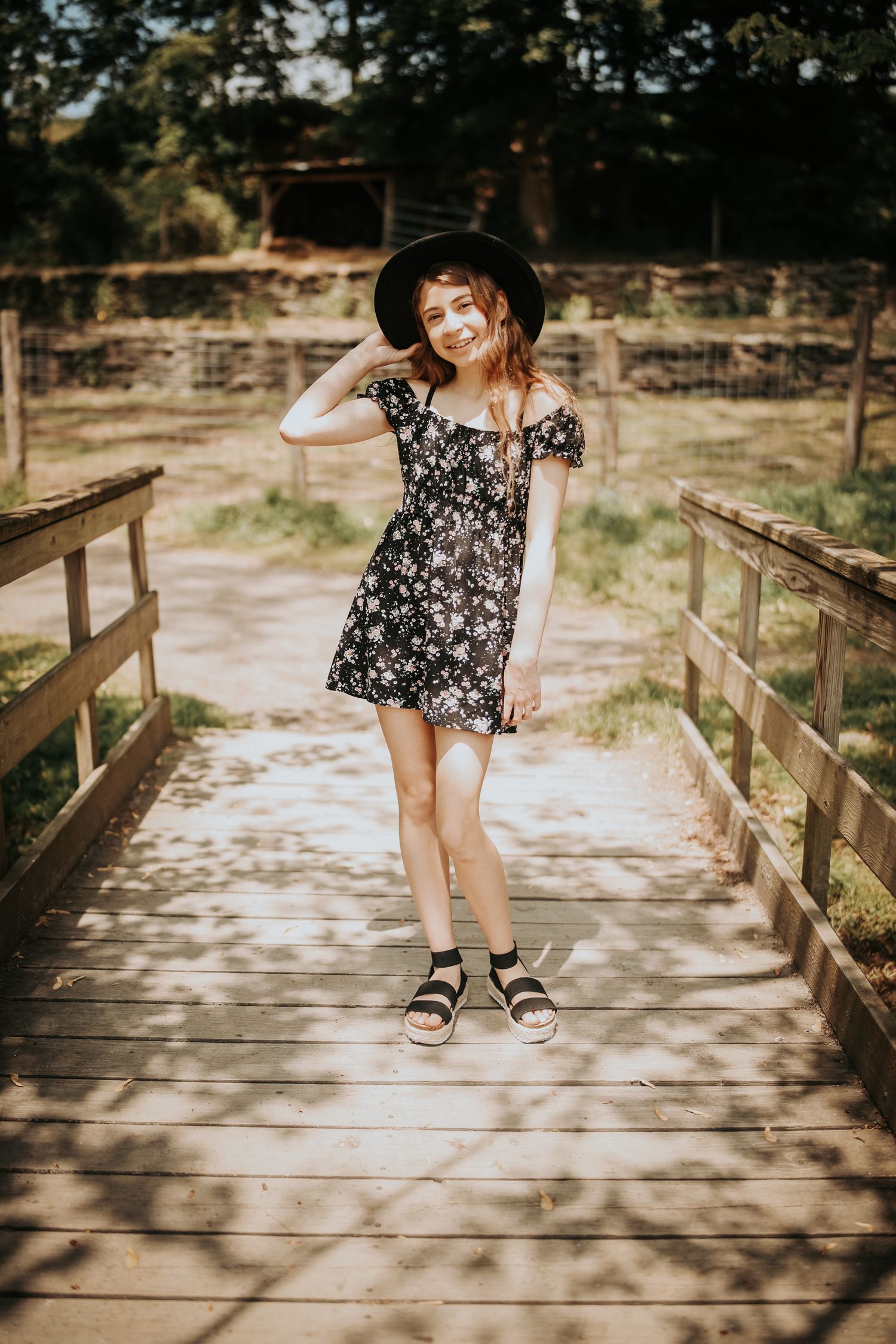 A floral dress and black hat create a summery look on a wooden bridge in a natural outdoor setting.