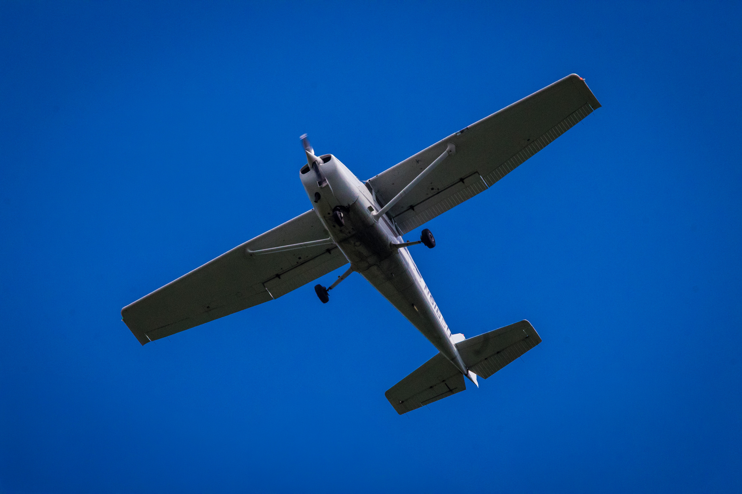 Small single-engine propeller plane flying overhead against a clear blue sky, viewed from below.