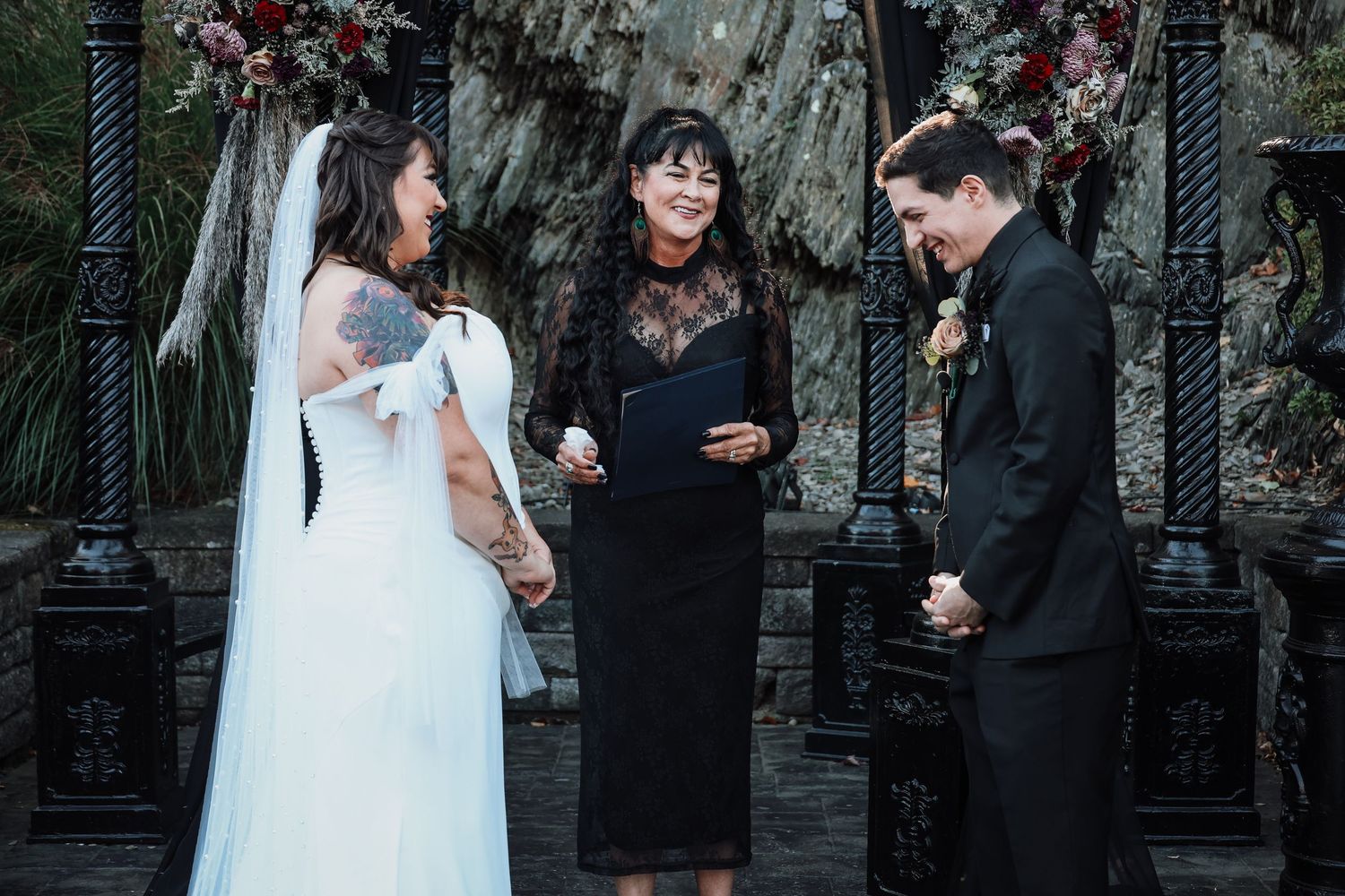 A wedding couple and officiant stand together during an outdoor ceremony with autumn foliage in the background.