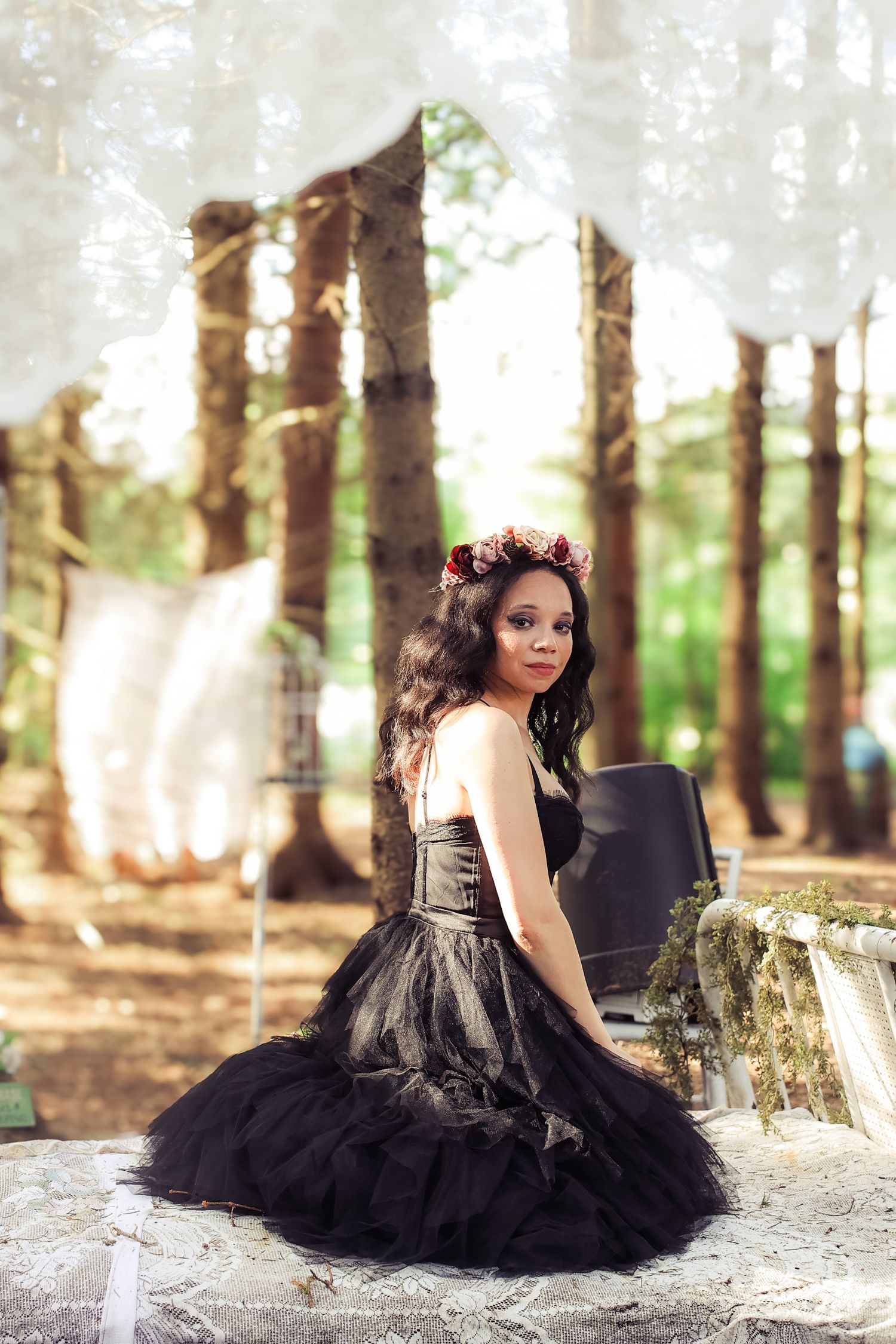 A girl in a black tulle dress poses gracefully in a forest setting with sunlight filtering through the trees.