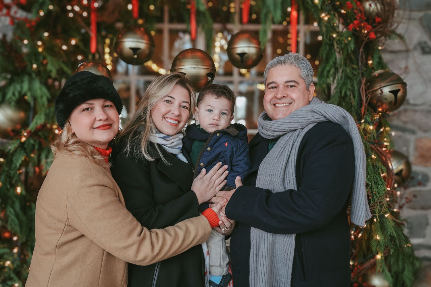 A group poses together in winter scarves and coats in front of a decorated Christmas tree with red ribbons and bells.