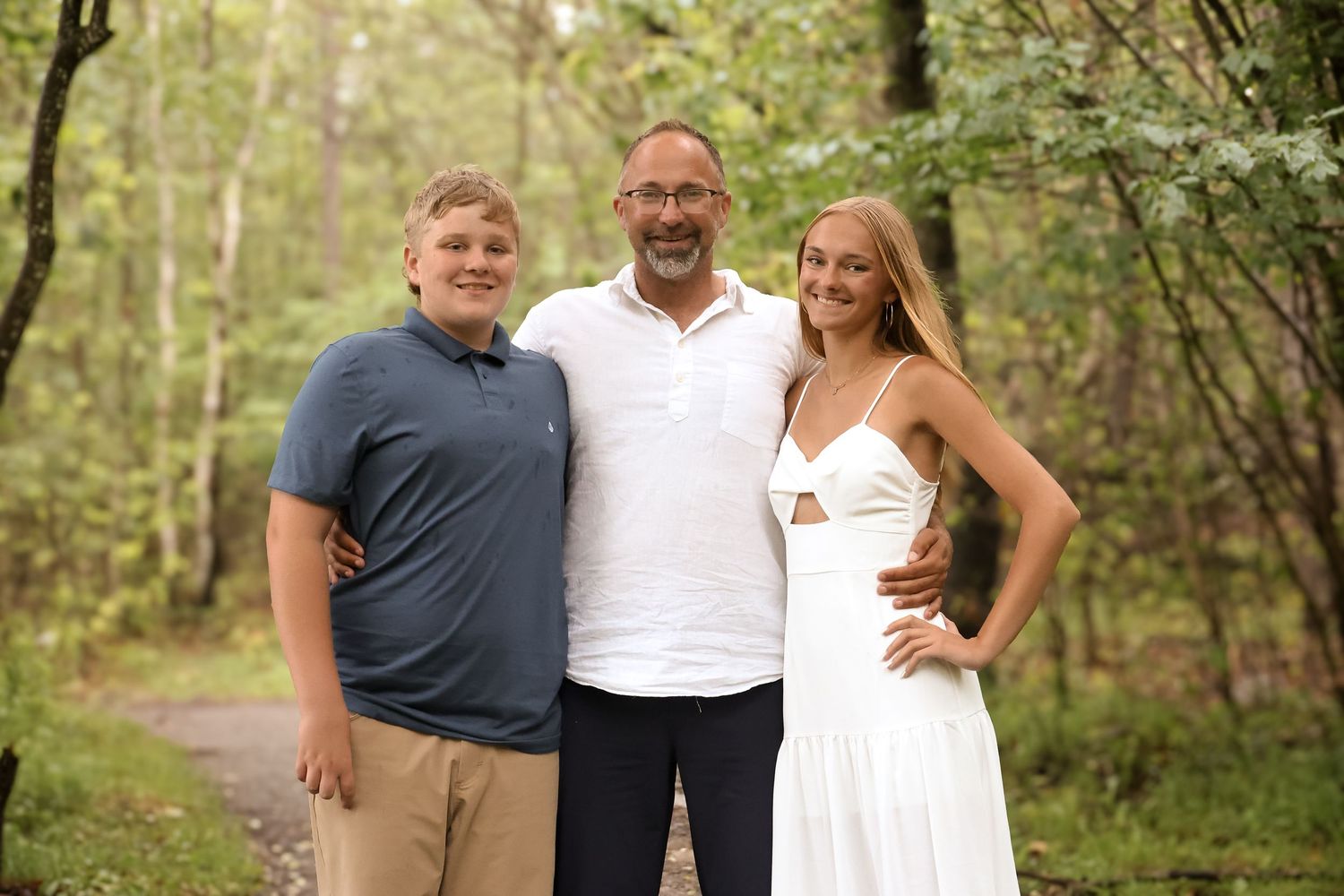 Three people stand together on a wooded path, with one wearing a white dress and others in casual attire.