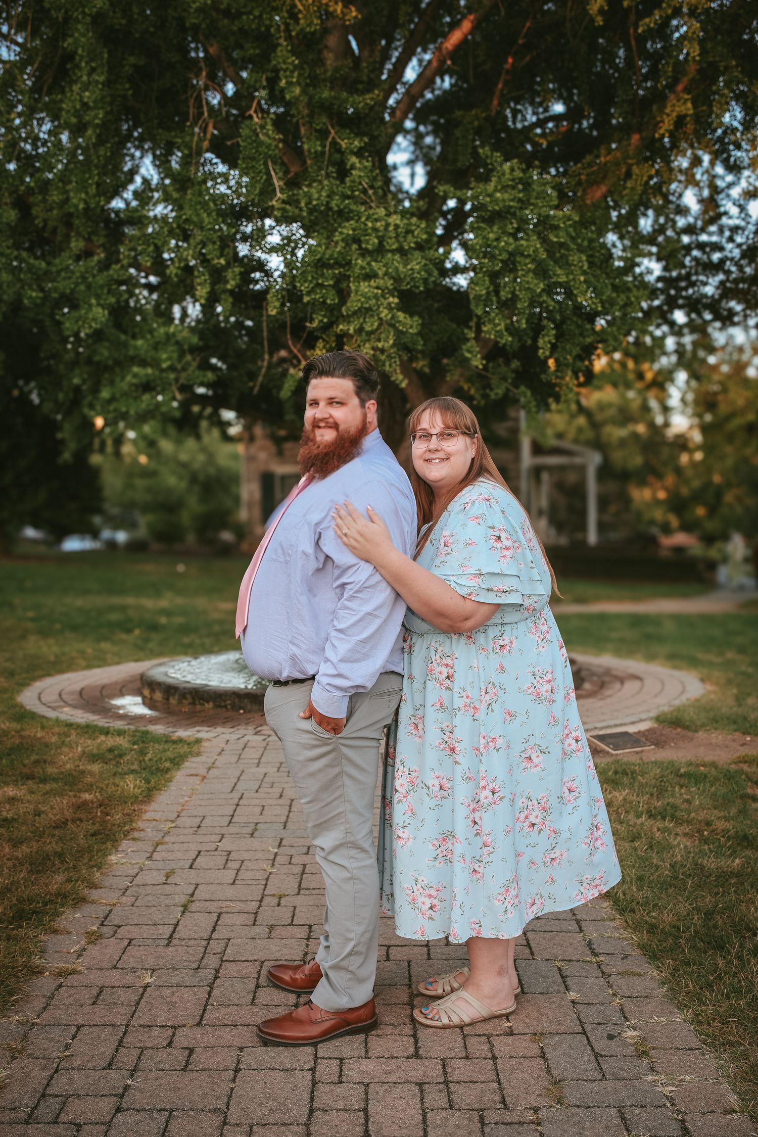 A couple stands together on a brick path in a garden setting during golden hour, with green trees in the background.