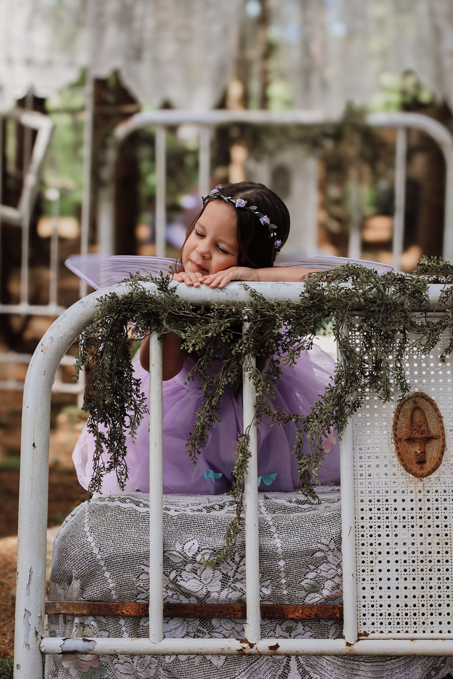 A decorative vintage white headboard adorned with purple fabric and Spanish moss creates a dreamy outdoor setting.