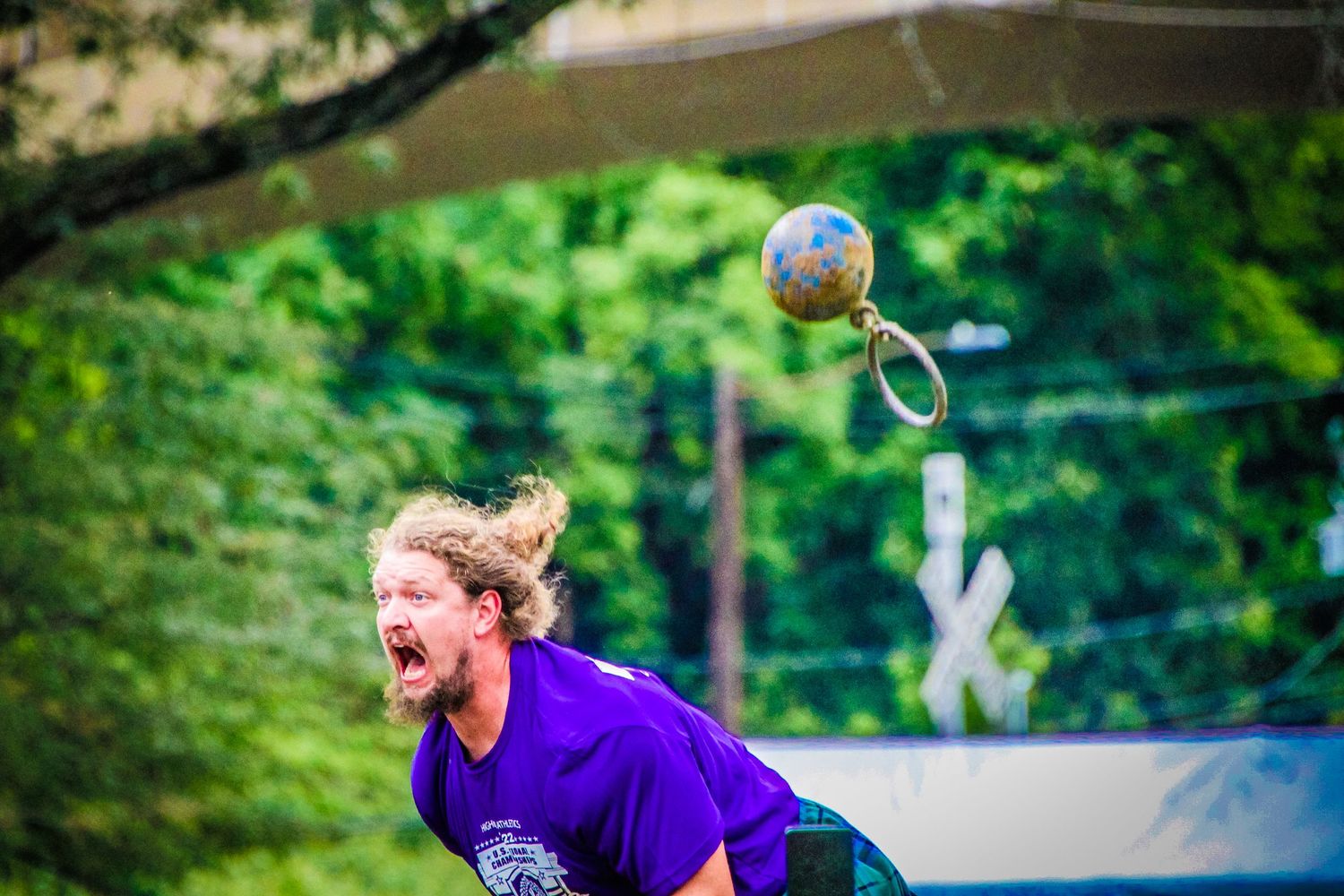 Player in purple jersey reacts during intense volleyball match with ball flying overhead in outdoor forest setting.