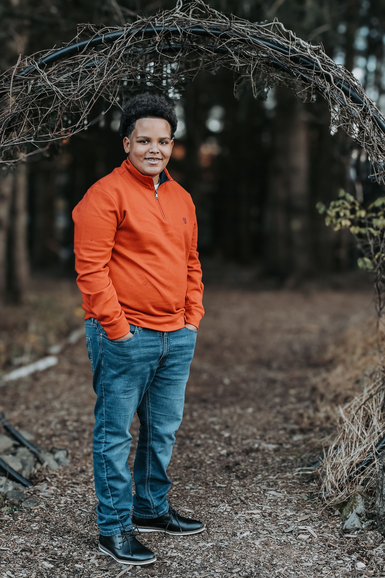 Person in bright orange sweater and blue jeans stands on forest trail in moody outdoor setting.