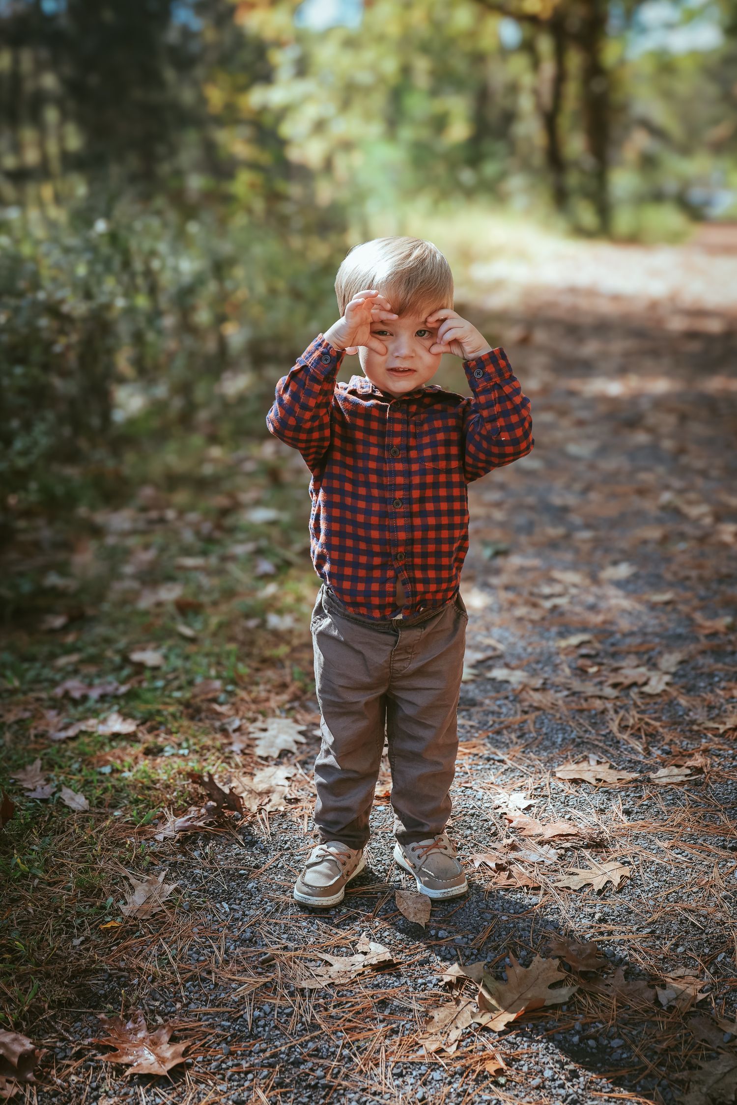 A small child in a red plaid shirt and brown pants stands on a forest path covered in fallen autumn leaves.