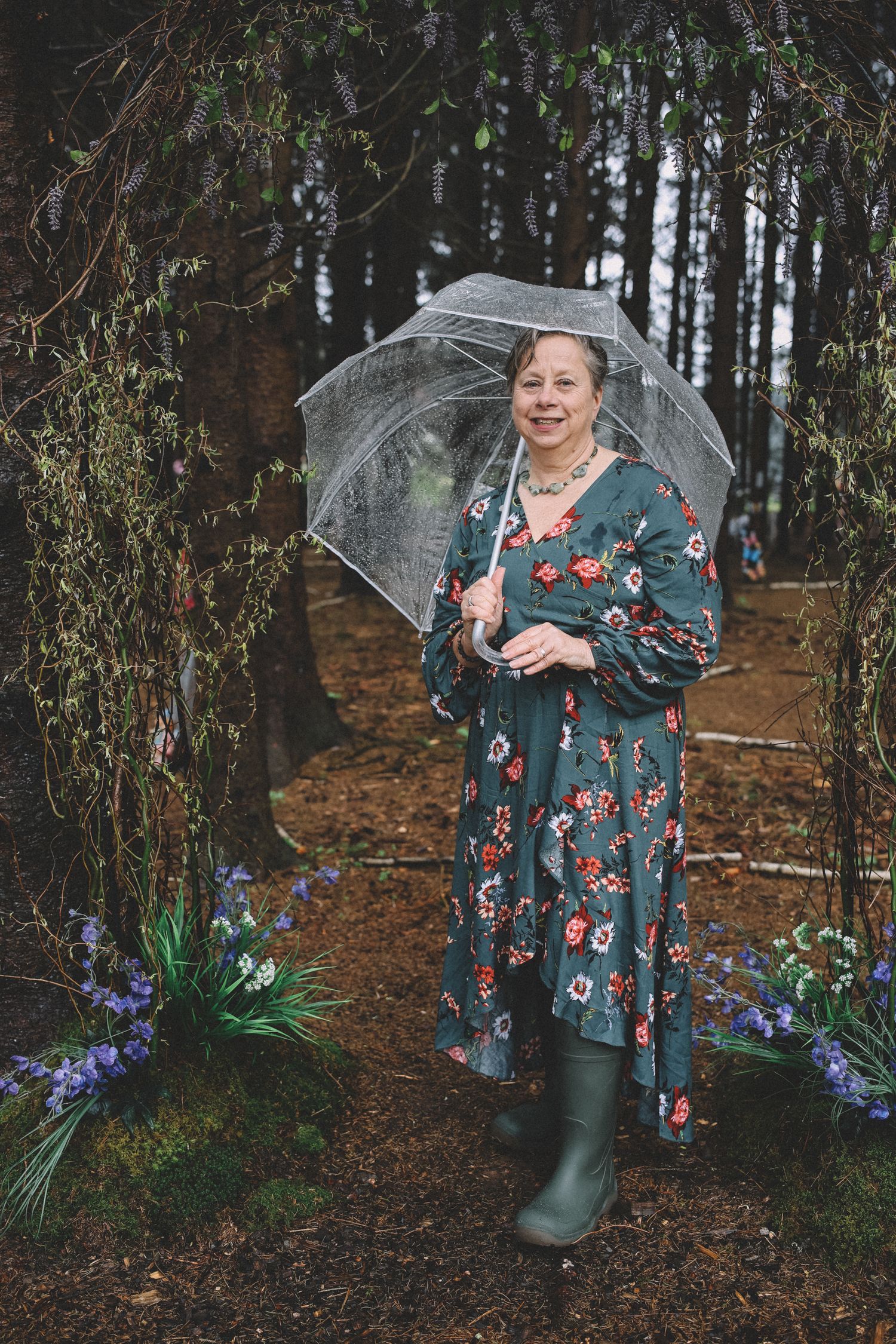 A person in a floral dress and rain boots poses with a clear umbrella in a moody forest setting.