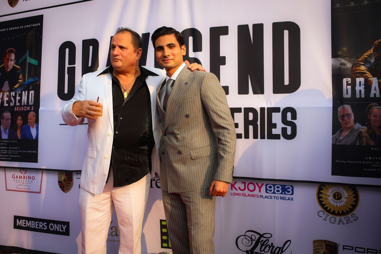 Two people pose together at a red carpet event with promotional banners visible in the background.