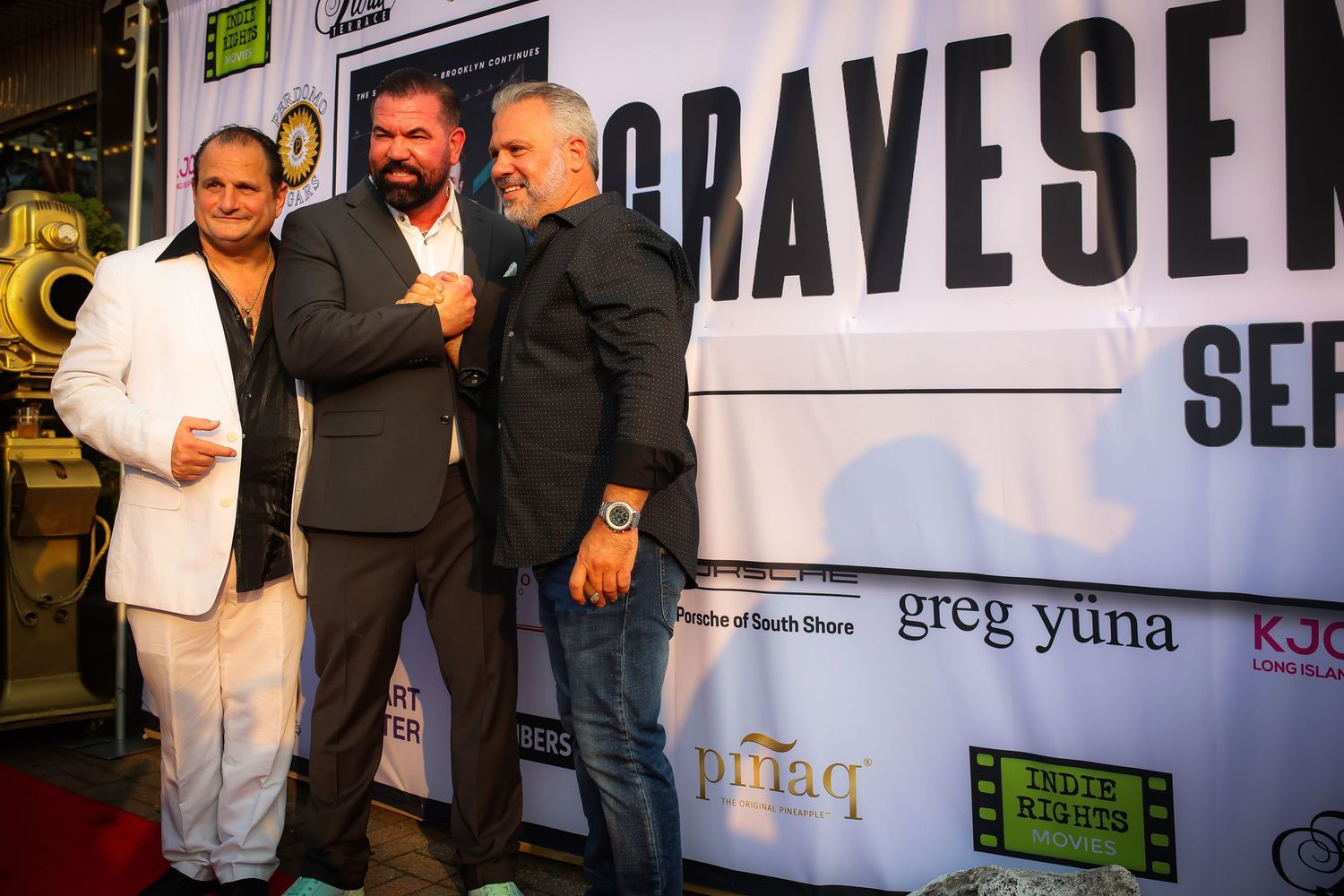 Three men in formal attire pose together at a media event with Bravese signage visible in the background.