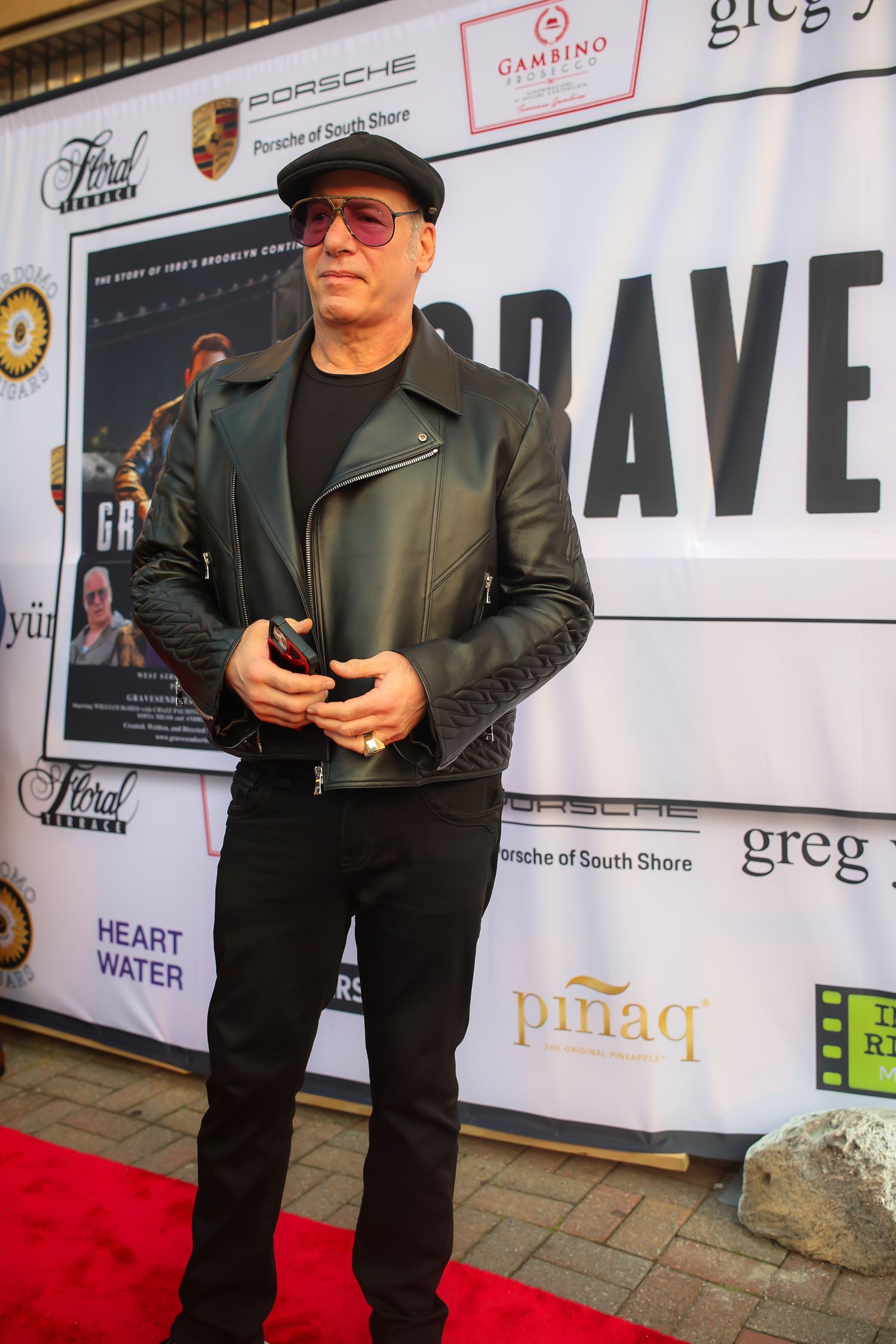 Person wearing black attire and glasses stands against event backdrop with sponsor logos and red carpet.