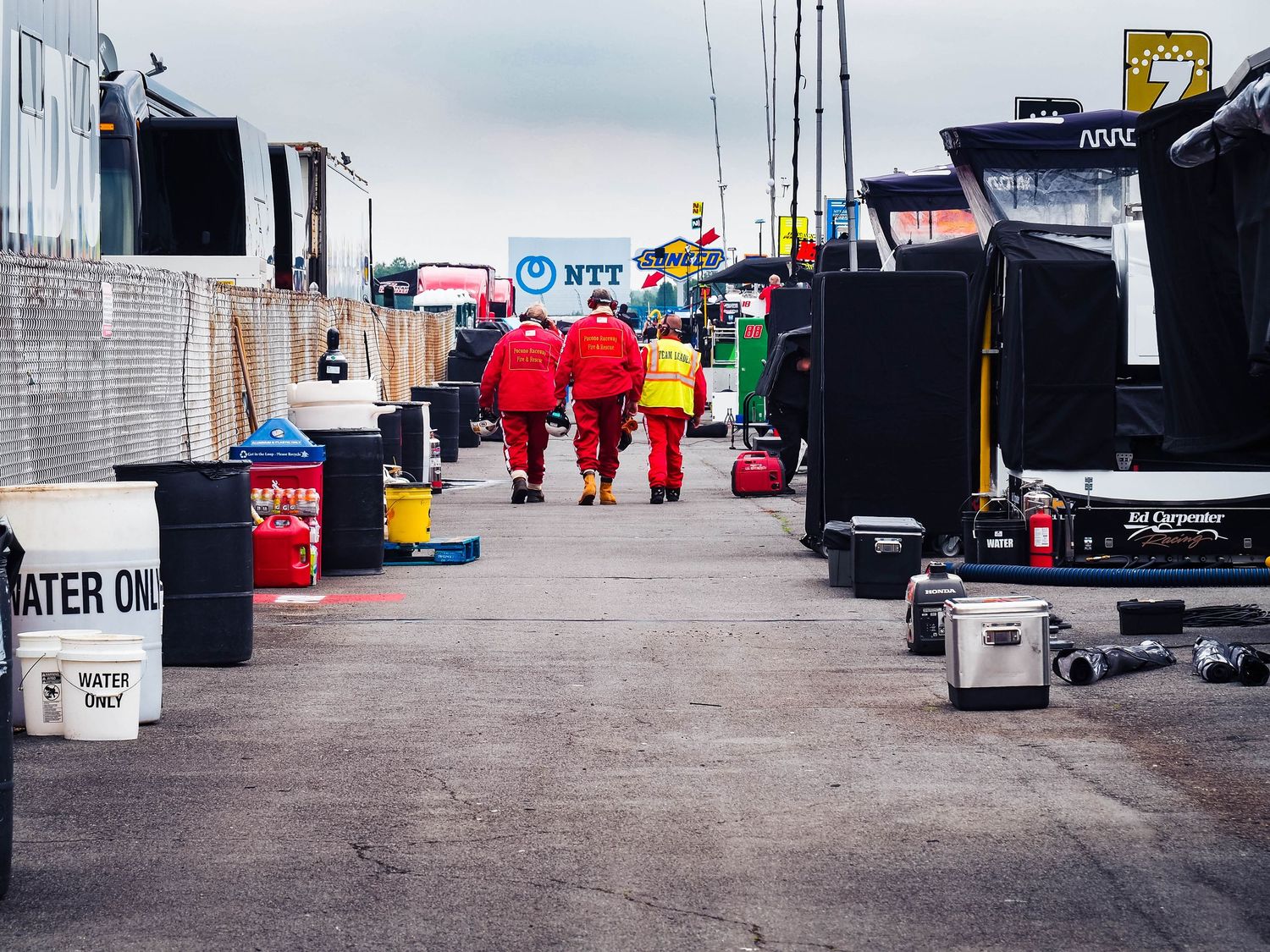 Racing team pit crew members work in a crowded garage area with equipment and tools during a motorsports event.