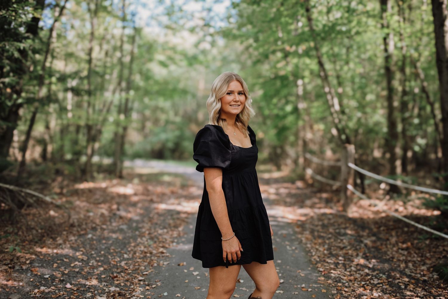Woman in black dress poses along a wooded path with natural lighting filtering through the trees.