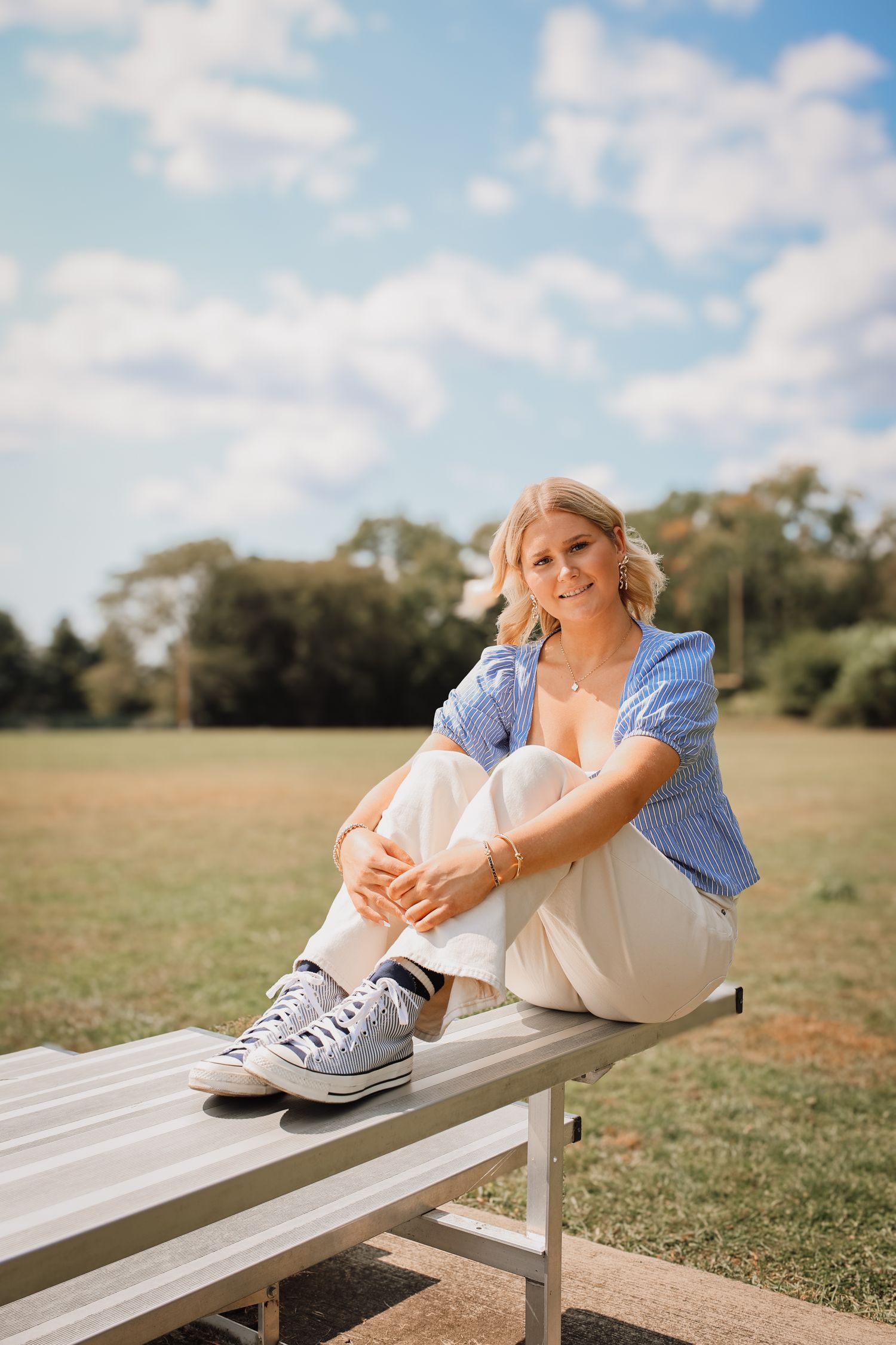 Woman in blue shirt and white pants relaxes on wooden bench in sunny outdoor setting.