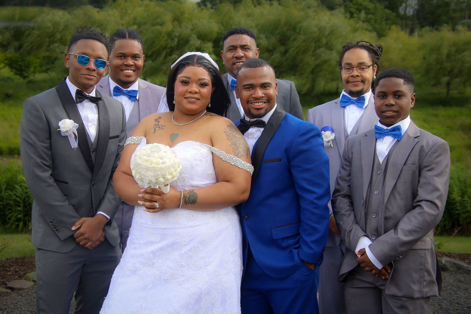 Wedding party members in blue and gray suits pose with the bride in a white gown during an outdoor celebration.