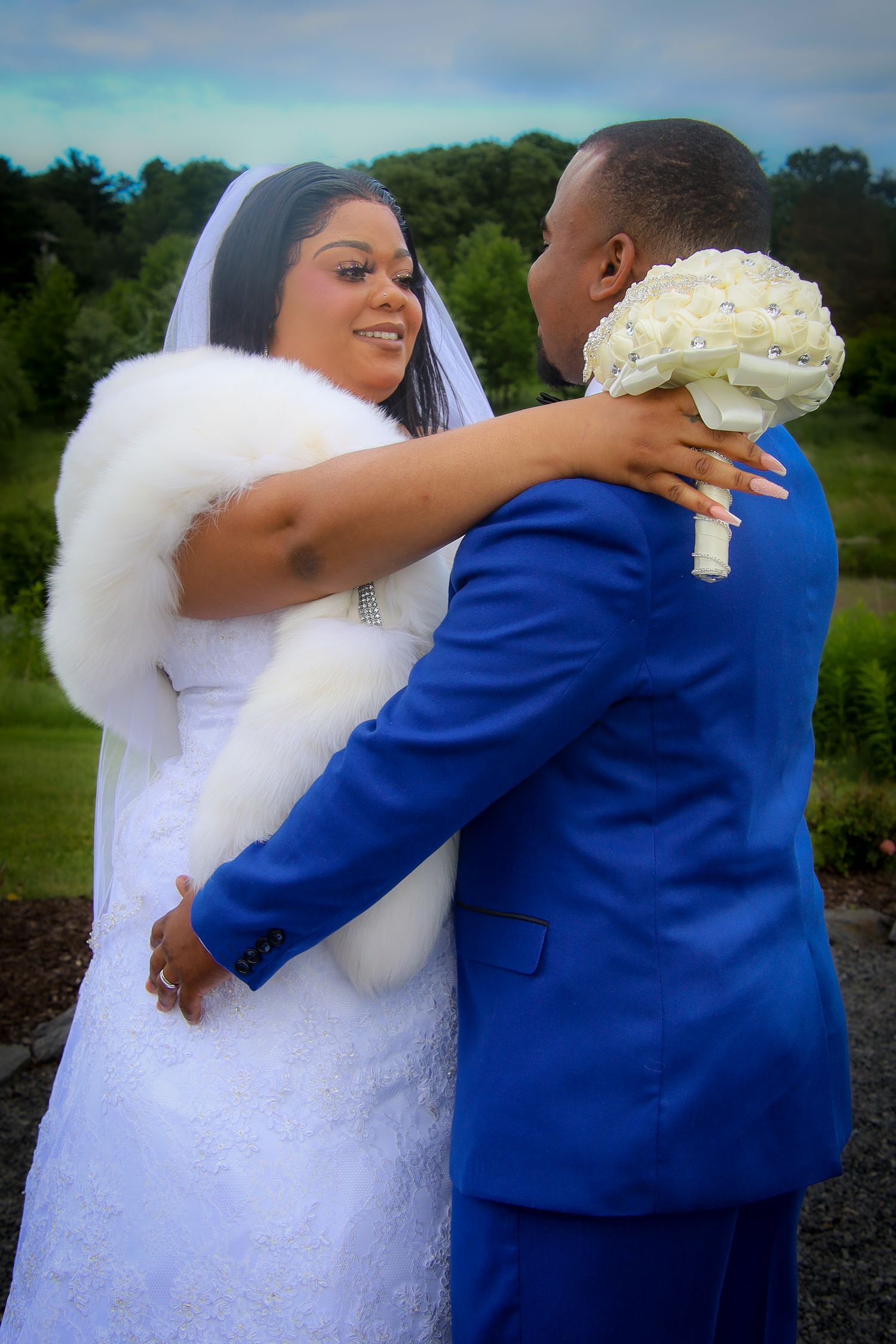 A bride in a white dress and fur wrap embraces someone wearing a royal blue suit while holding a white bouquet outdoors.