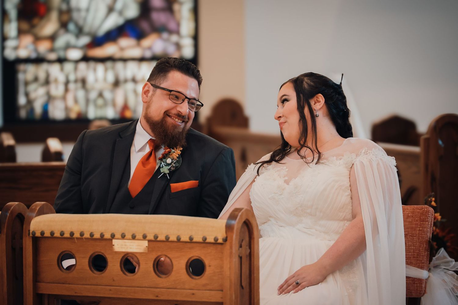 A couple shares a tender moment while sitting together in a wooden church pew during their wedding ceremony.